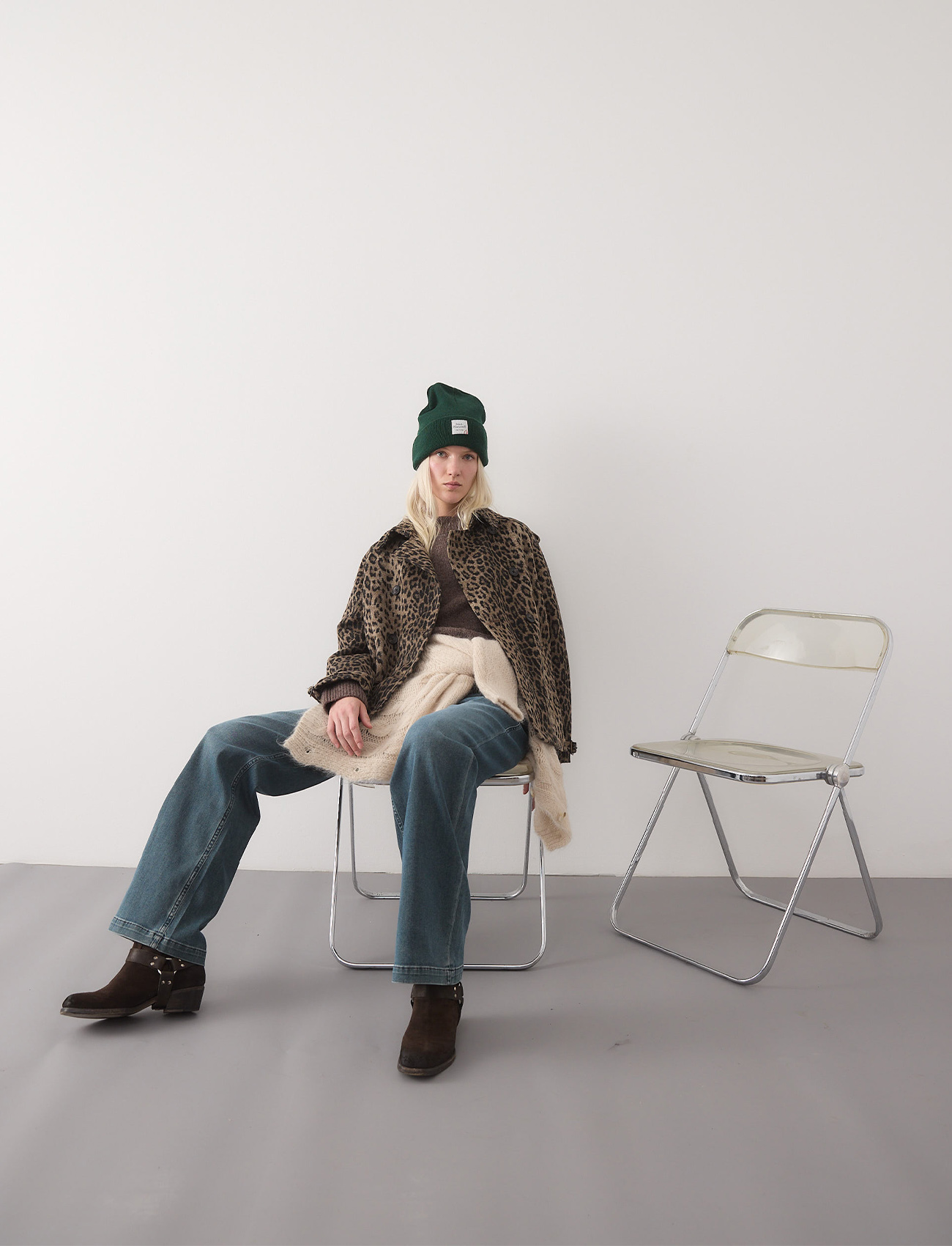 Eine Person, die eine grüne Mütze, eine Jacke mit Leopardenmuster, einen dunklen Pullover, blaue Jeans und braune Stiefel trägt, sitzt lässig auf einem Klappstuhl neben einem leeren Stuhl vor einer schlichten hellen Wand und einem grauen Boden.