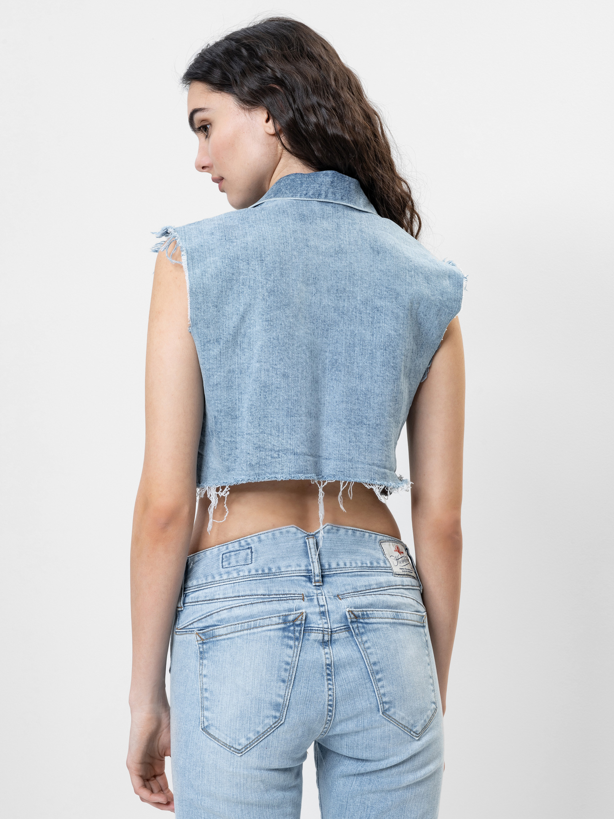 Eine Frau mit langen, gewellten dunklen Haaren wird von hinten gezeigt. Sie trägt eine ärmellose, ausgefranste Jeansweste und hellblaue, hochgekrempelte Jeans vor einem schlichten weißen Hintergrund.
