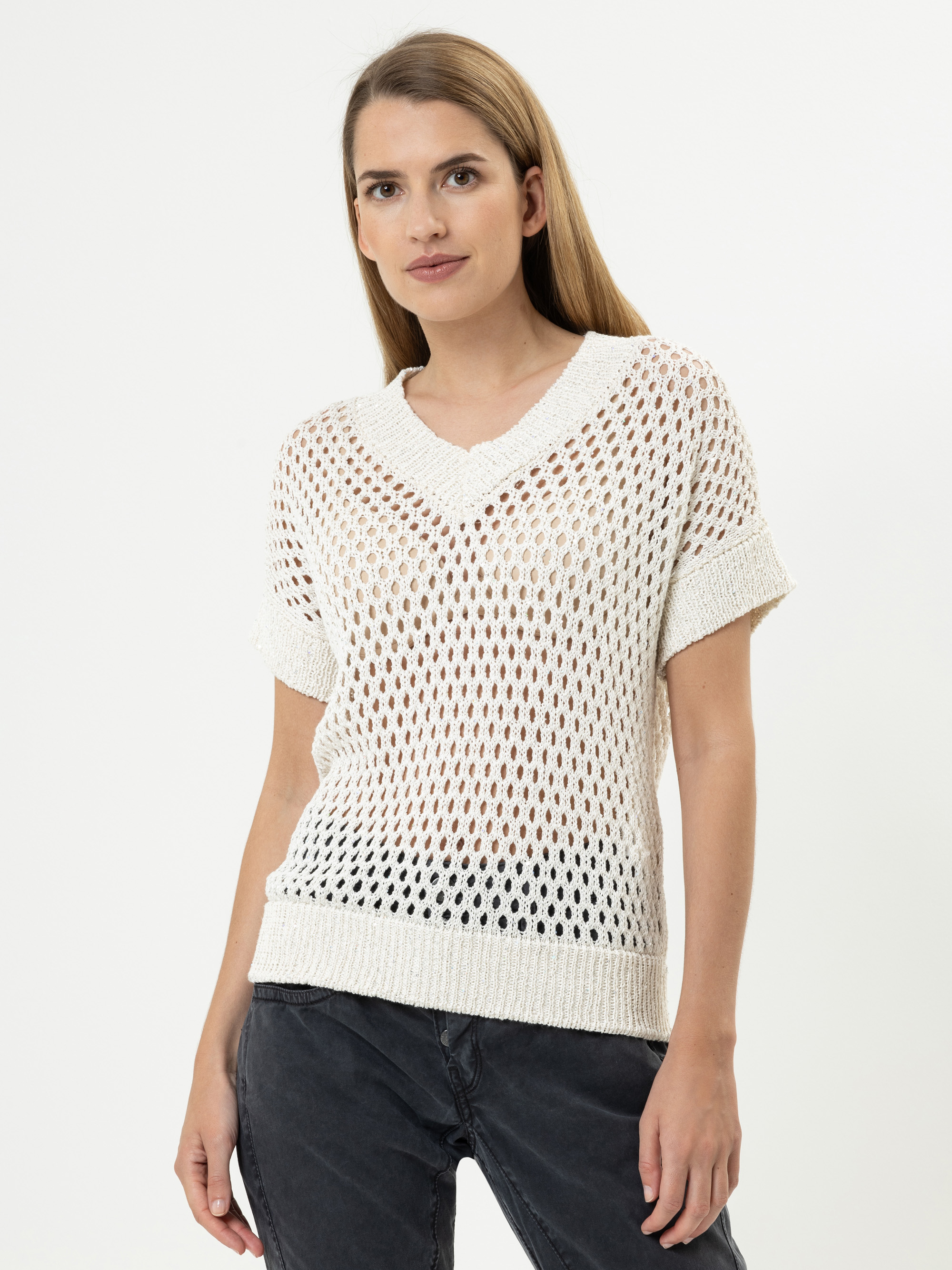 Eine Frau mit langem, glattem Haar trägt einen cremefarbenen, kurzärmeligen, offen gestrickten Pullover über einem dunklen Oberteil und einer dunklen Jeans, die vor einem schlichten weißen Hintergrund steht.