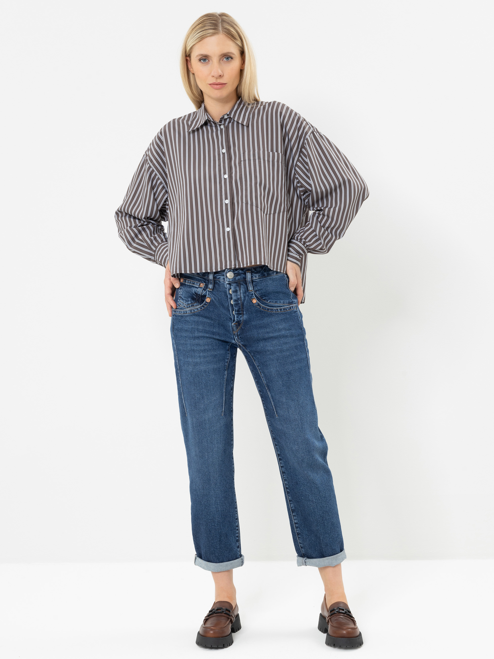 Vor einem weißen Hintergrund steht eine Frau, die ein grau-weiß gestreiftes Hemd, eine blaue Jeans mit Umschlag und braune Slipper trägt. Sie hat blondes Haar und steckt die Hände in die Taschen.