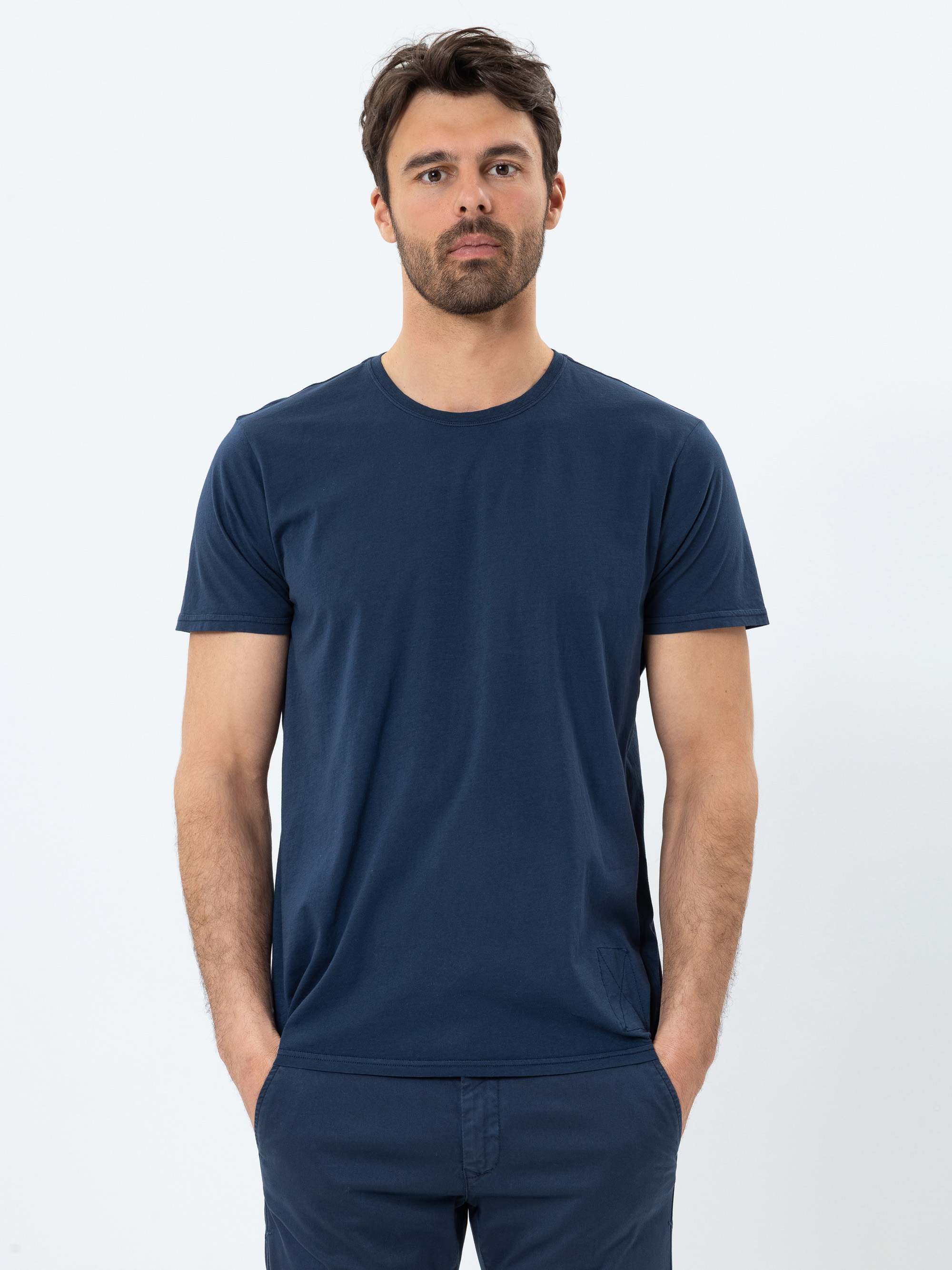 Ein Mann mit kurzen braunen Haaren und Bart steht vor einem schlichten weißen Hintergrund, trägt ein marineblaues T-Shirt und eine passende Hose und hat die Hände in den Taschen.