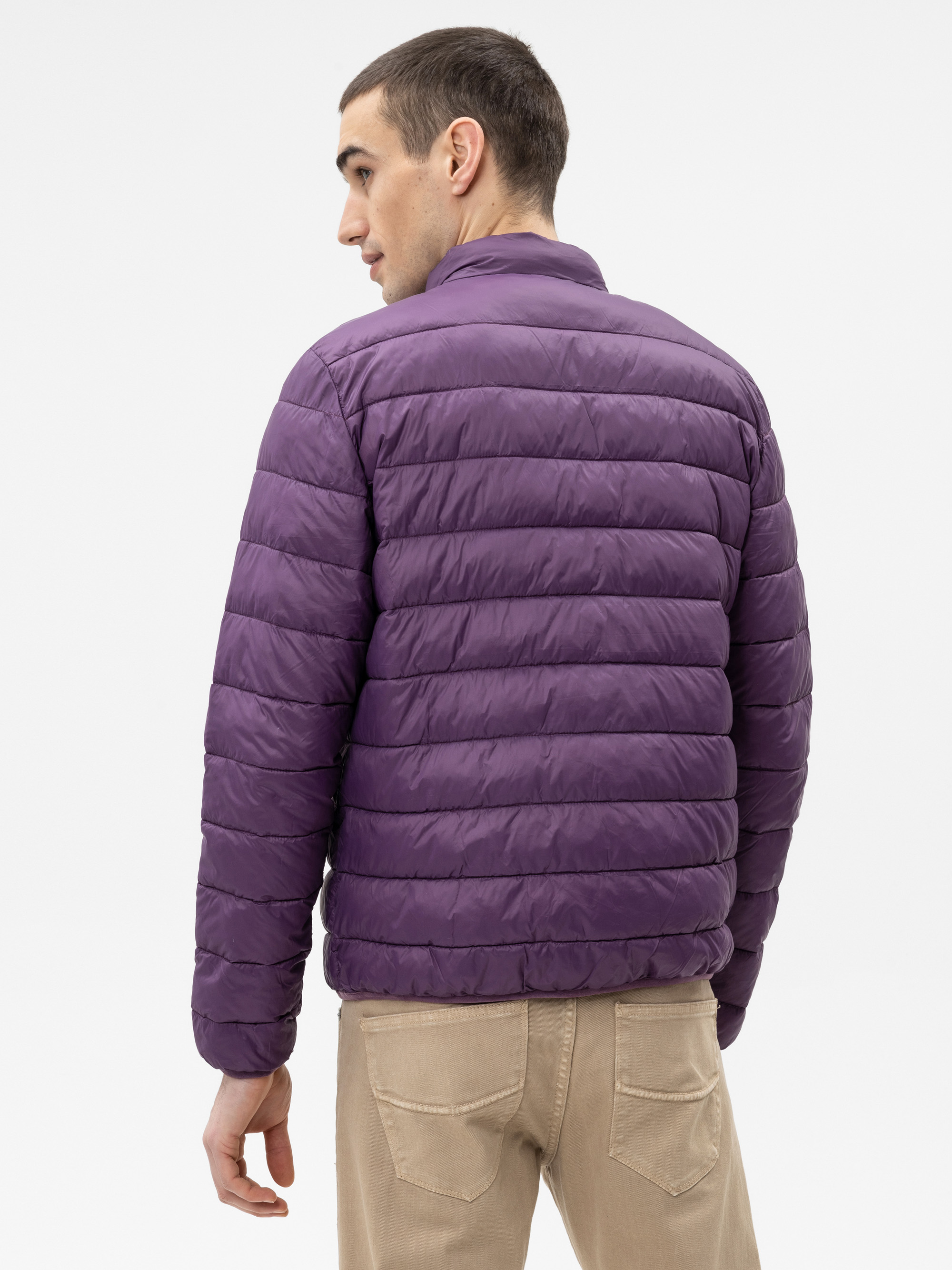 Ein Mann steht mit dem Gesicht zur Wand, trägt eine gesteppte lila Pufferjacke und eine beigefarbene Hose vor einem schlichten weißen Hintergrund.