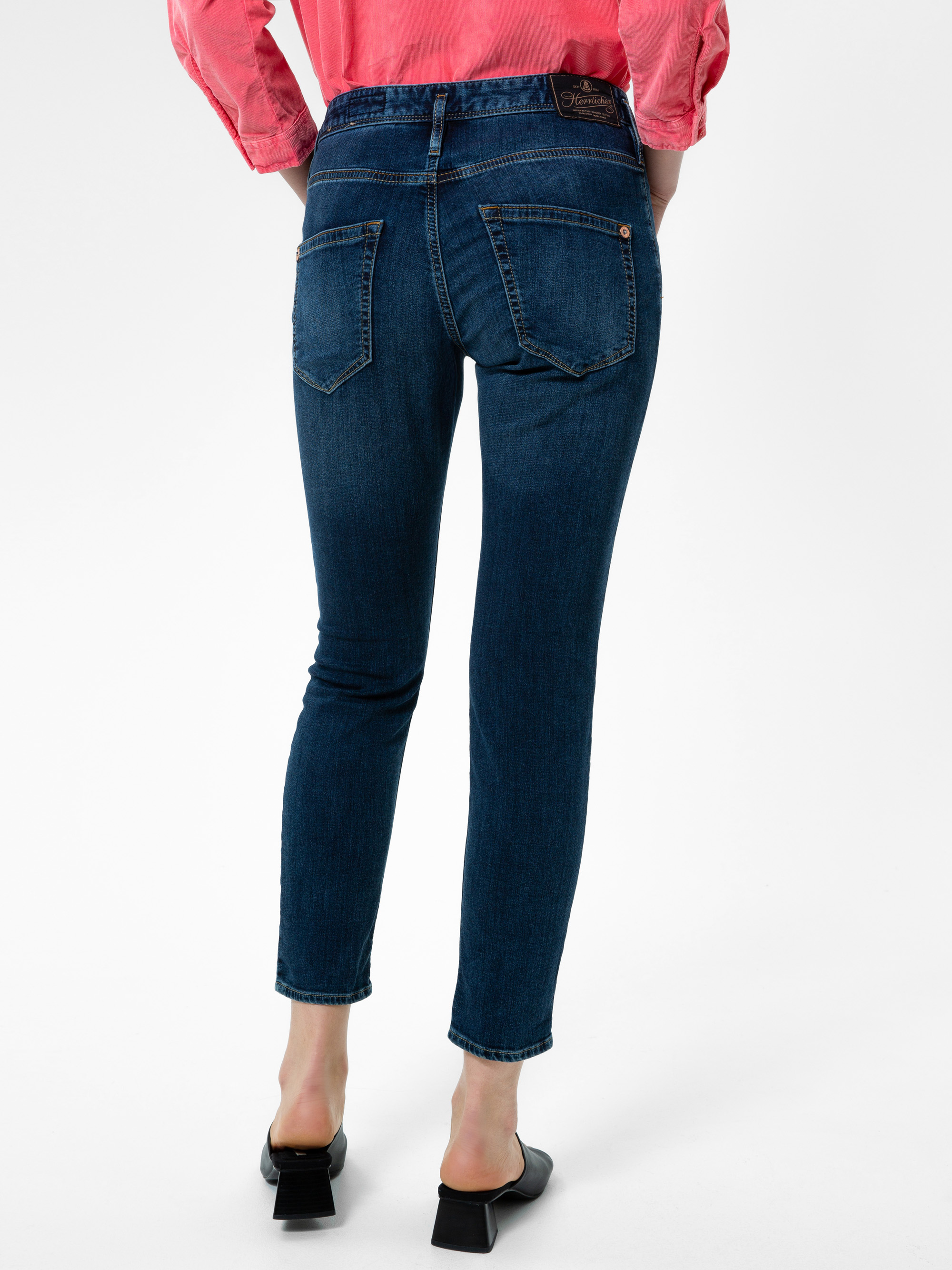 Person, die ein rosafarbenes langärmeliges Hemd, dunkelblaue Röhrenjeans und schwarze Schuhe mit Absätzen trägt, von hinten vor einem schlichten weißen Hintergrund.