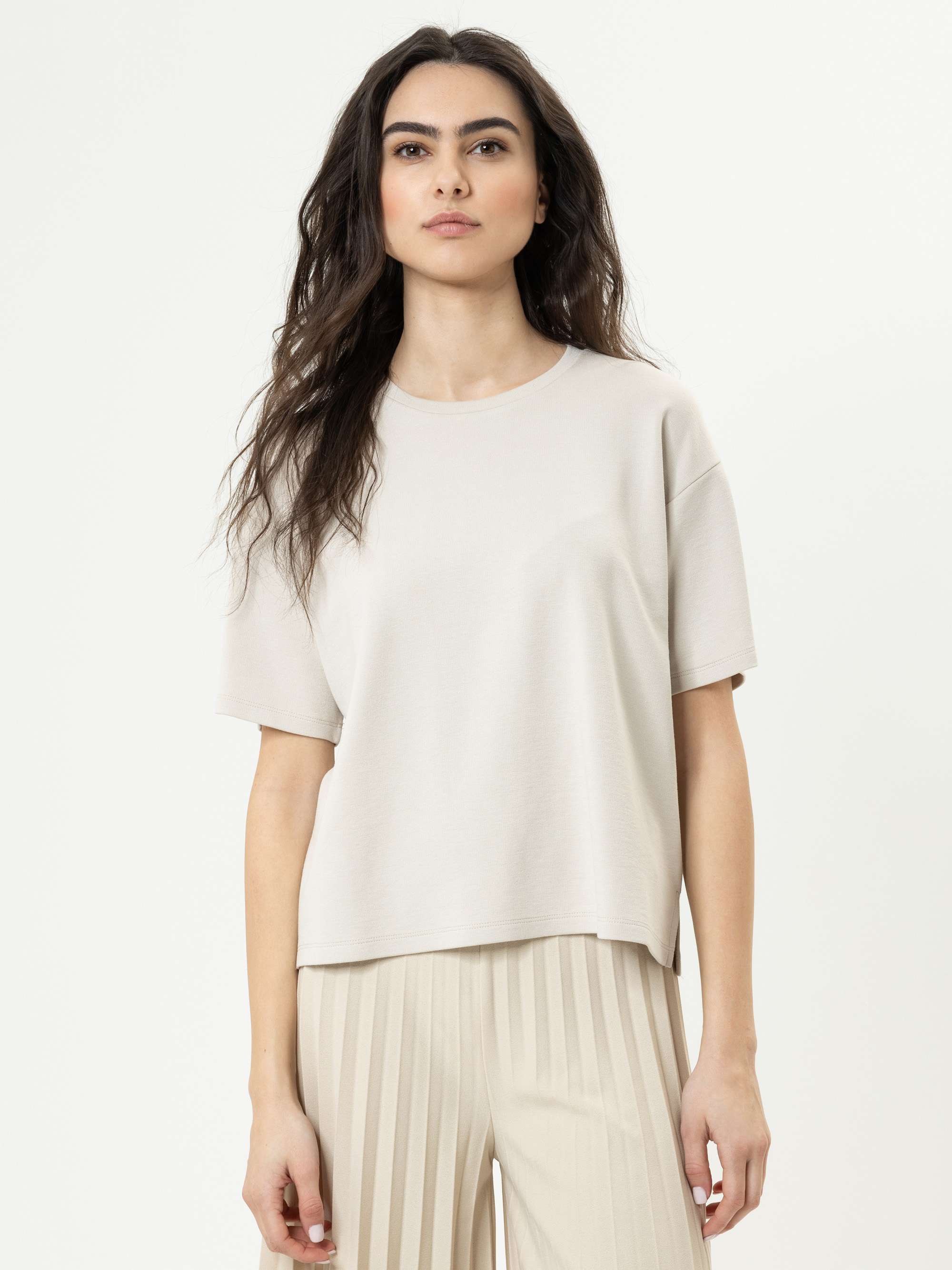 Eine Frau mit langen dunklen Haaren trägt ein locker sitzendes beiges T-Shirt und eine hellbeige gestreifte Hose. Sie steht vor einem schlichten weißen Hintergrund und schaut mit neutralem Blick direkt in die Kamera.