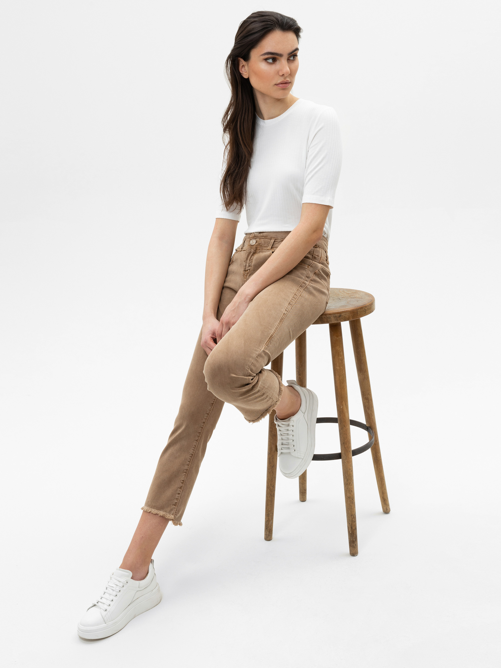 Eine Frau mit langen dunklen Haaren, die ein weißes kurzärmeliges Oberteil, eine hellbraune Hose und weiße Turnschuhe trägt, sitzt seitlich auf einem Holzhocker vor einem schlichten weißen Hintergrund.