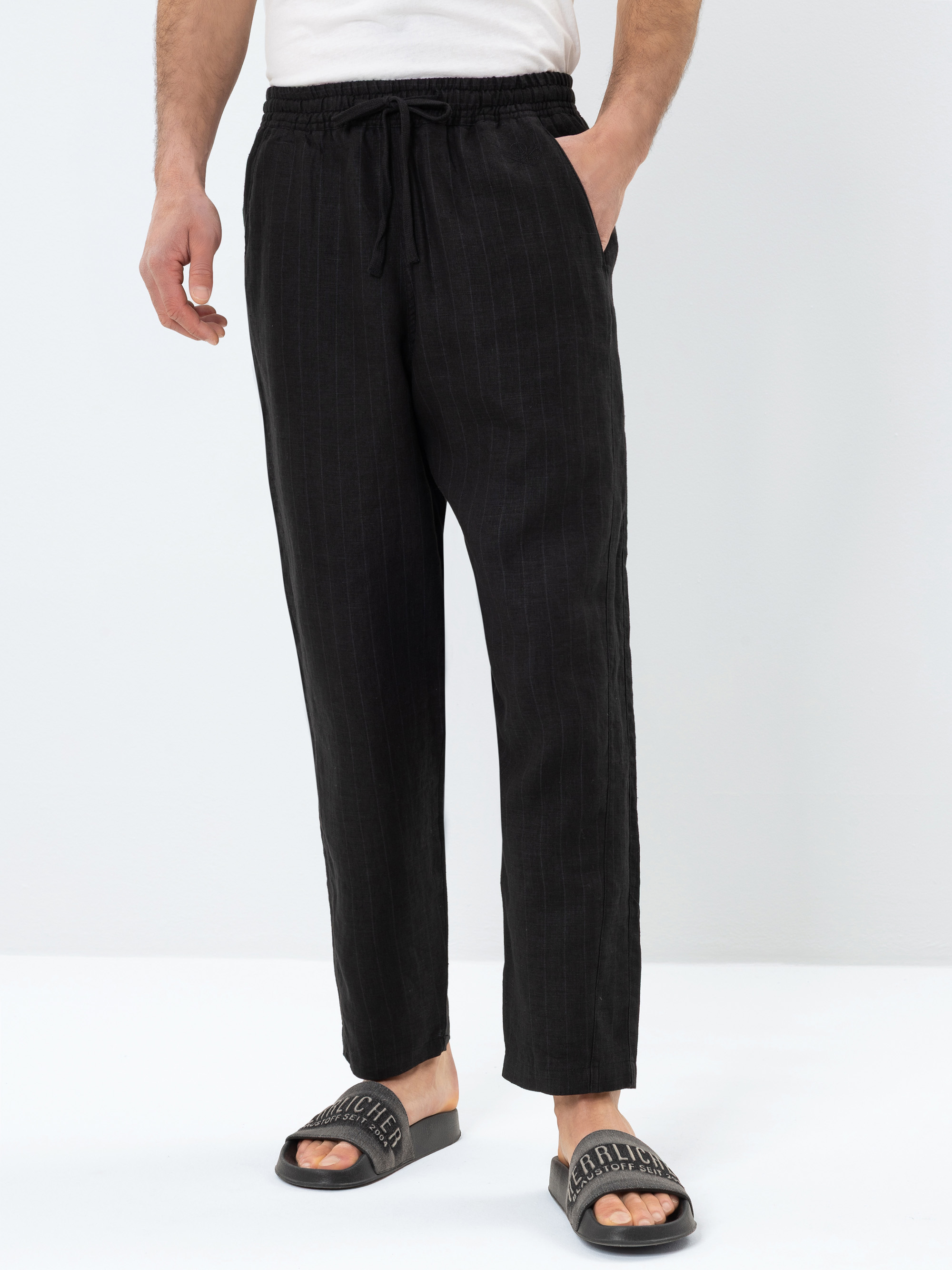 Eine Person, die eine schwarze Hose mit Nadelstreifen, ein weißes Hemd und schwarze Slide-Sandalen mit Aufschrift trägt, steht vor einem schlichten weißen Hintergrund.