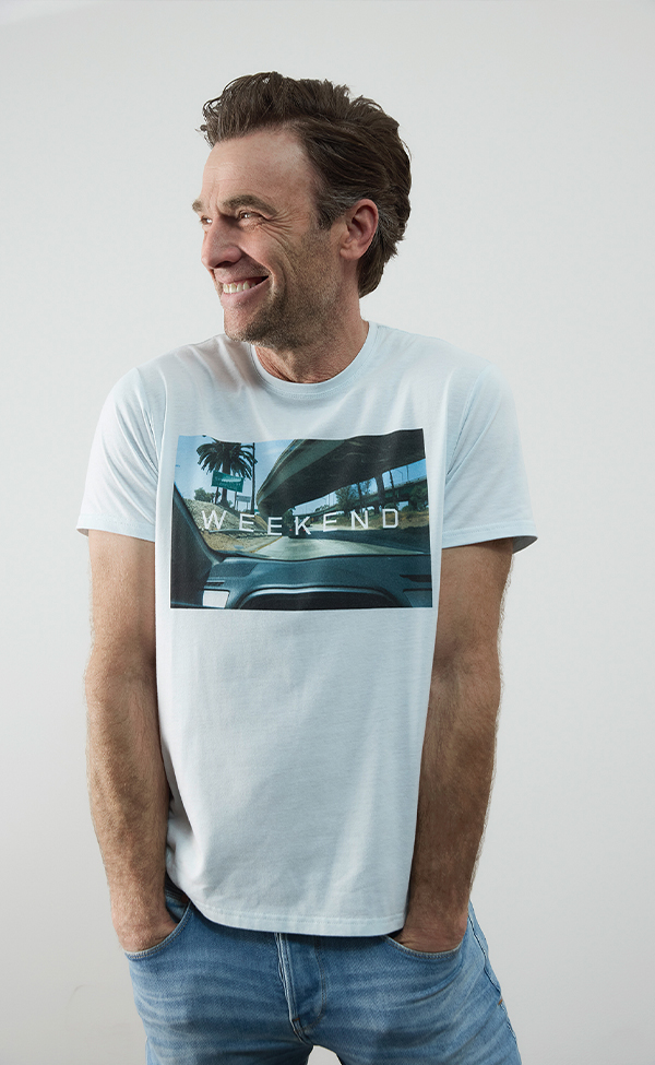 Ein Mann mit kurzen braunen Haaren lächelt und schaut dabei nach links. Er trägt ein hellblaues T-Shirt mit dem Aufdruck „WEEKEND“ und eine hellblaue Jeans. Der Hintergrund ist einfarbig und hell.