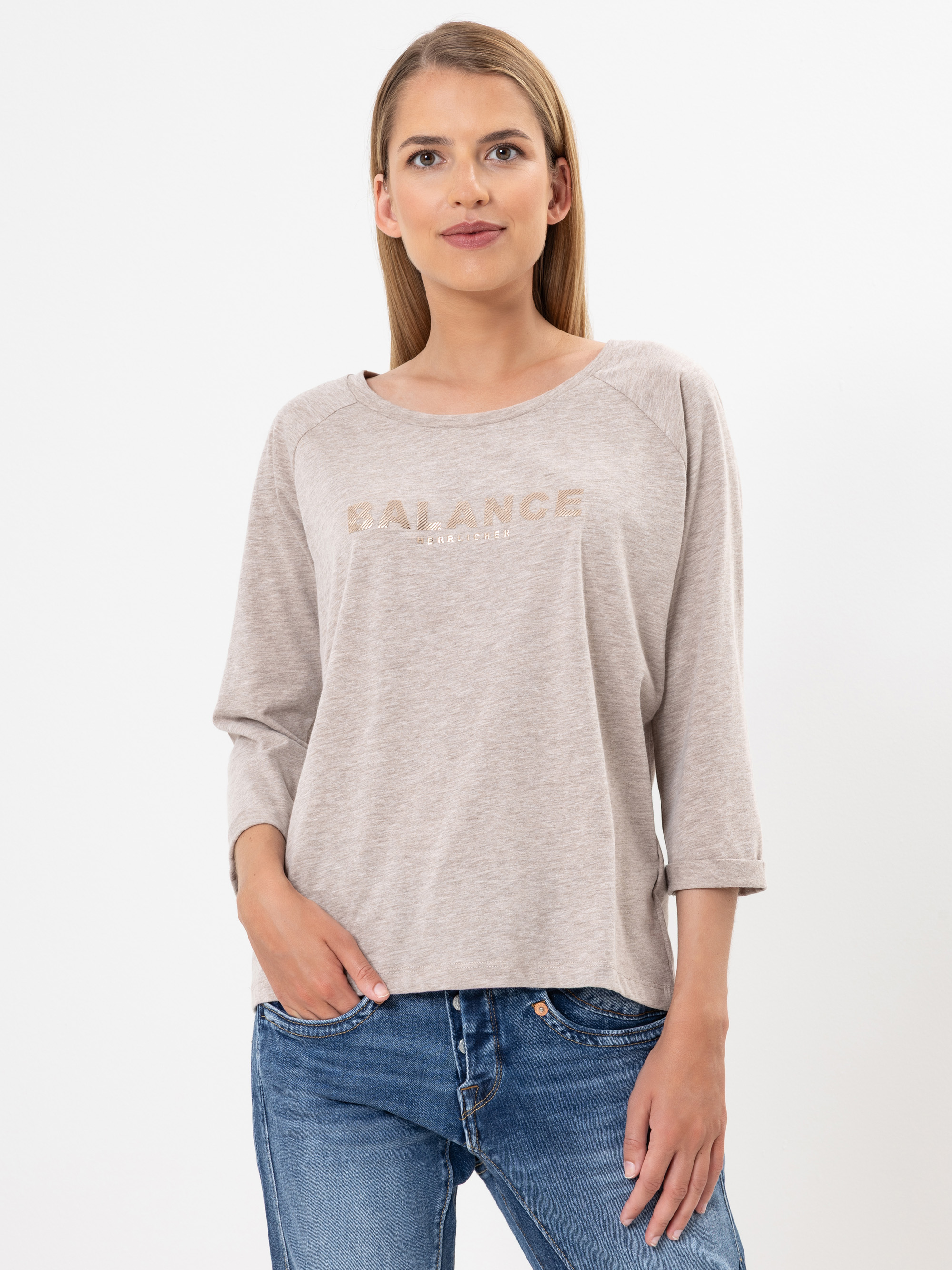 Eine Frau mit glattem, hellbraunem Haar trägt ein beiges, langärmeliges Hemd mit dem Aufdruck BALANCE auf der Vorderseite und blaue Jeans. Sie steht vor einem schlichten weißen Hintergrund, hat eine Hand in der Tasche und lächelt leicht.