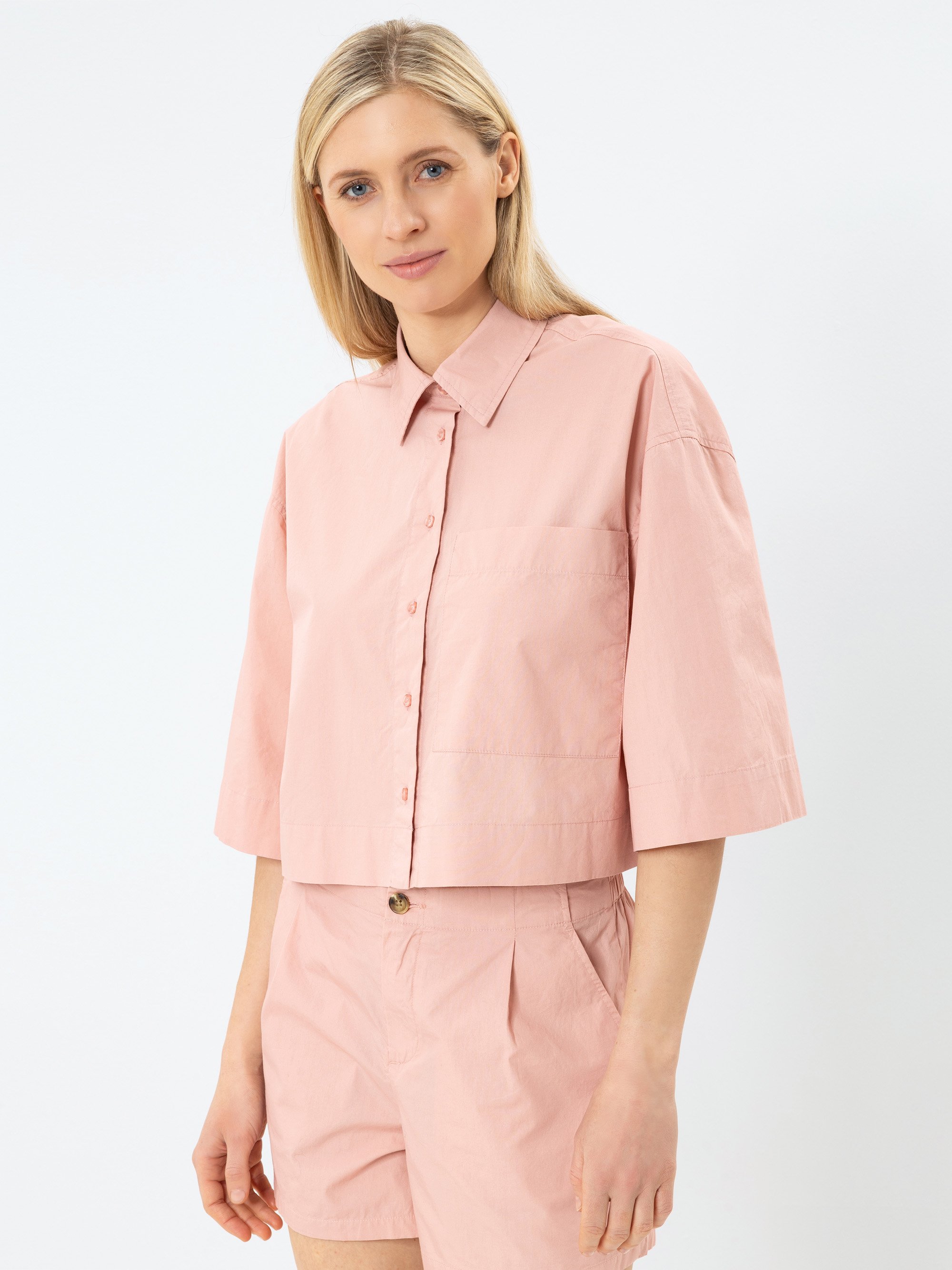 Eine Frau mit langen blonden Haaren trägt ein hellrosa, kurzärmeliges Button-up-Shirt und dazu passende hochgeschnittene Shorts und steht vor einem schlichten weißen Hintergrund.