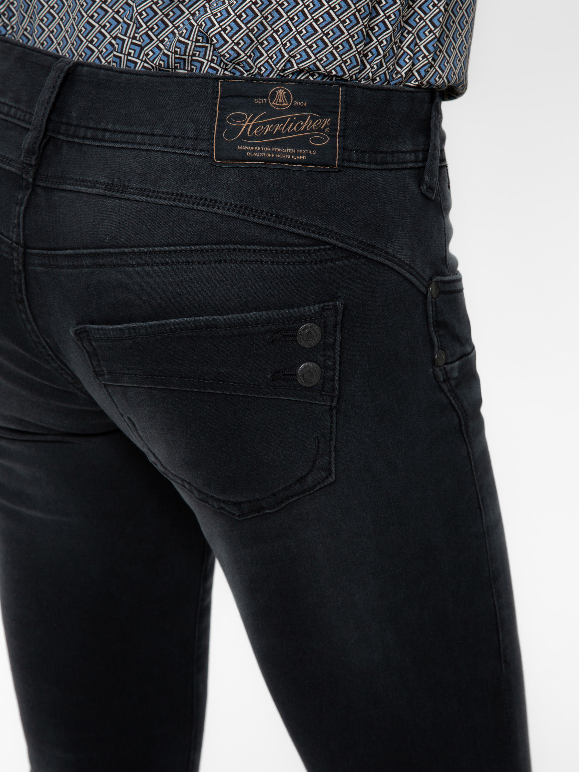 Nahaufnahme des Rückens einer Person, die eine dunkle Denim-Jeans mit einem einzigartigen Taschenmuster und einem braunen Lederetikett der Marke Herrlicher am Bund trägt. Die Person trägt außerdem ein gemustertes Hemd, das in die Hose gesteckt wird.