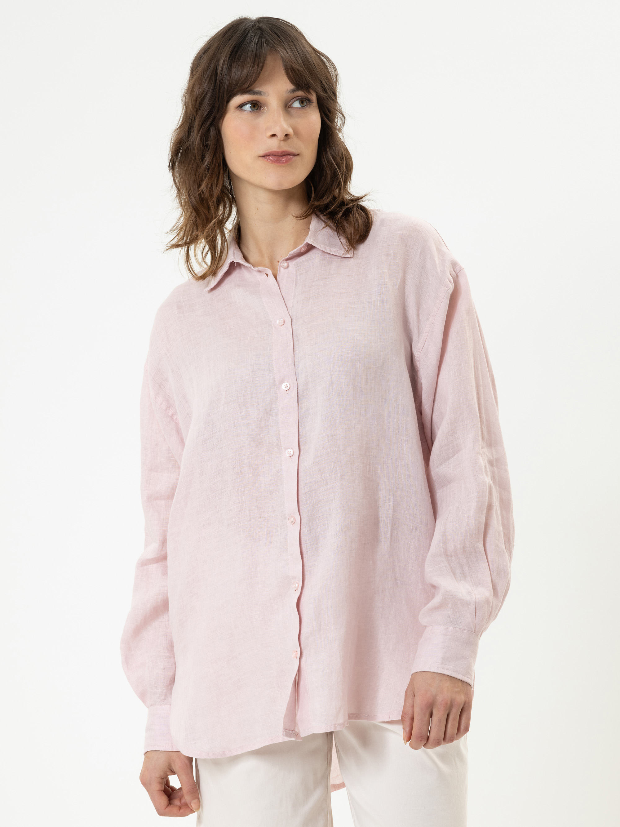 Eine Frau mit gewelltem braunem Haar trägt ein lockeres, hellrosafarbenes Button-up-Hemd und eine weiße Hose. Sie steht vor einem schlichten weißen Hintergrund und blickt mit einem entspannten Gesichtsausdruck leicht zur Seite.