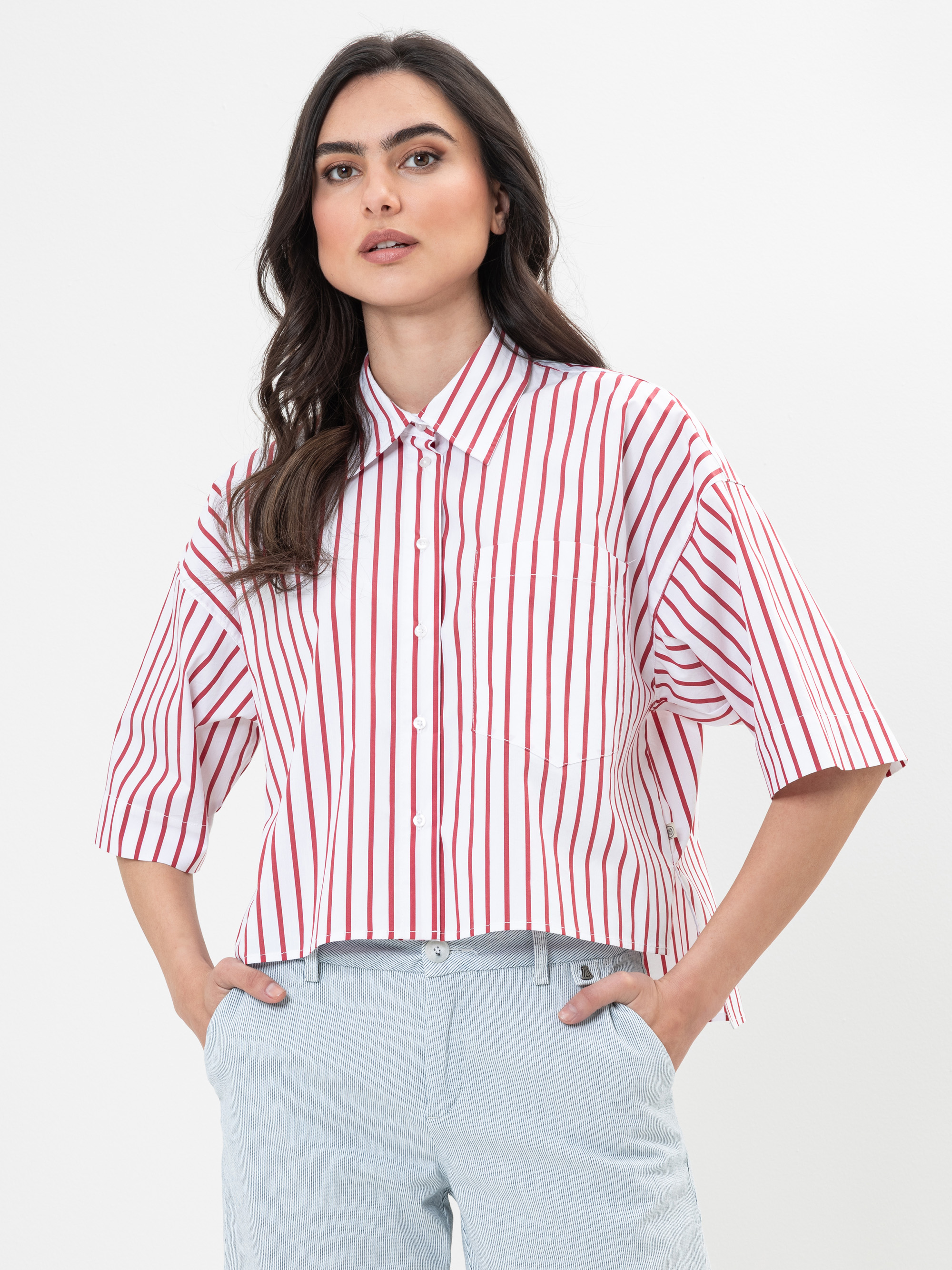 Eine Frau in einem rot-weiß vertikal gestreiften Button-up-Hemd mit ellbogenlangen Ärmeln und einer hellgrauen Hose steht vor einem schlichten weißen Hintergrund und blickt selbstbewusst in die Kamera.
