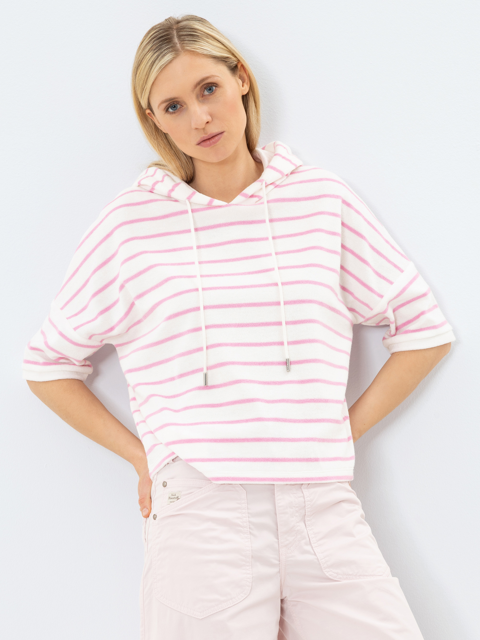 Eine Frau mit blonden Haaren, die einen weißen Kapuzenpullover mit rosa Querstreifen und eine hellrosa Hose trägt, steht vor einem weißen Hintergrund und schaut mit neutralem Blick in die Kamera.