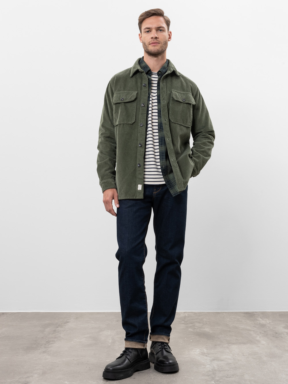 Ein Mann steht vor einem schlichten weißen Hintergrund und trägt eine grüne Cordjacke über einem gestreiften Hemd, dunkle Jeans und graue Schnürschuhe. Eine Hand steckt in seiner Tasche und er hat einen neutralen Gesichtsausdruck.