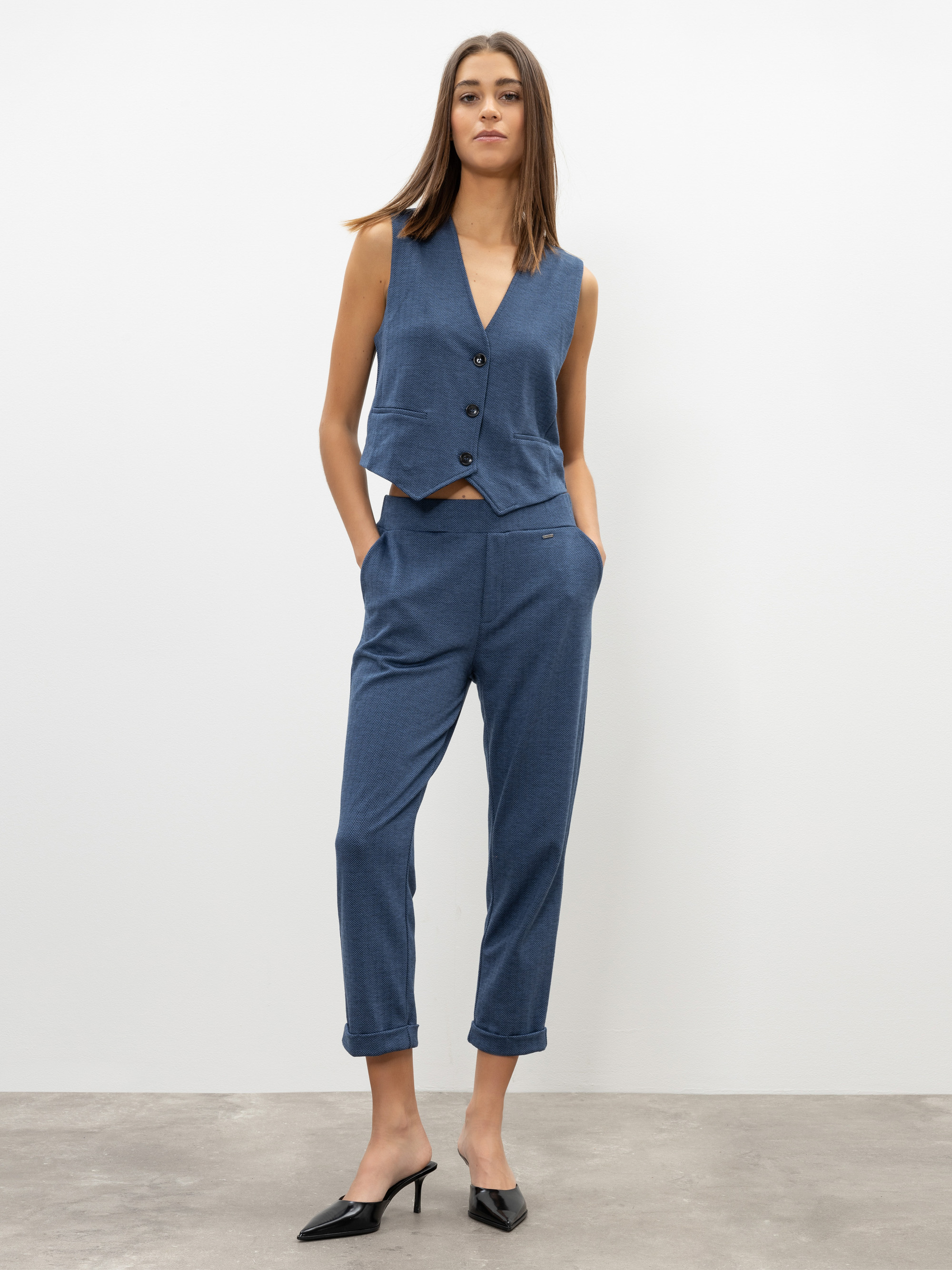 Eine Frau steht vor einem schlichten weißen Hintergrund und trägt eine ärmellose blaue Weste mit Knöpfen, eine dazu passende blaue Hose mit aufgerollten Manschetten und schwarze Pantoletten mit Absätzen. Sie hat die Hände in den Taschen und blickt n