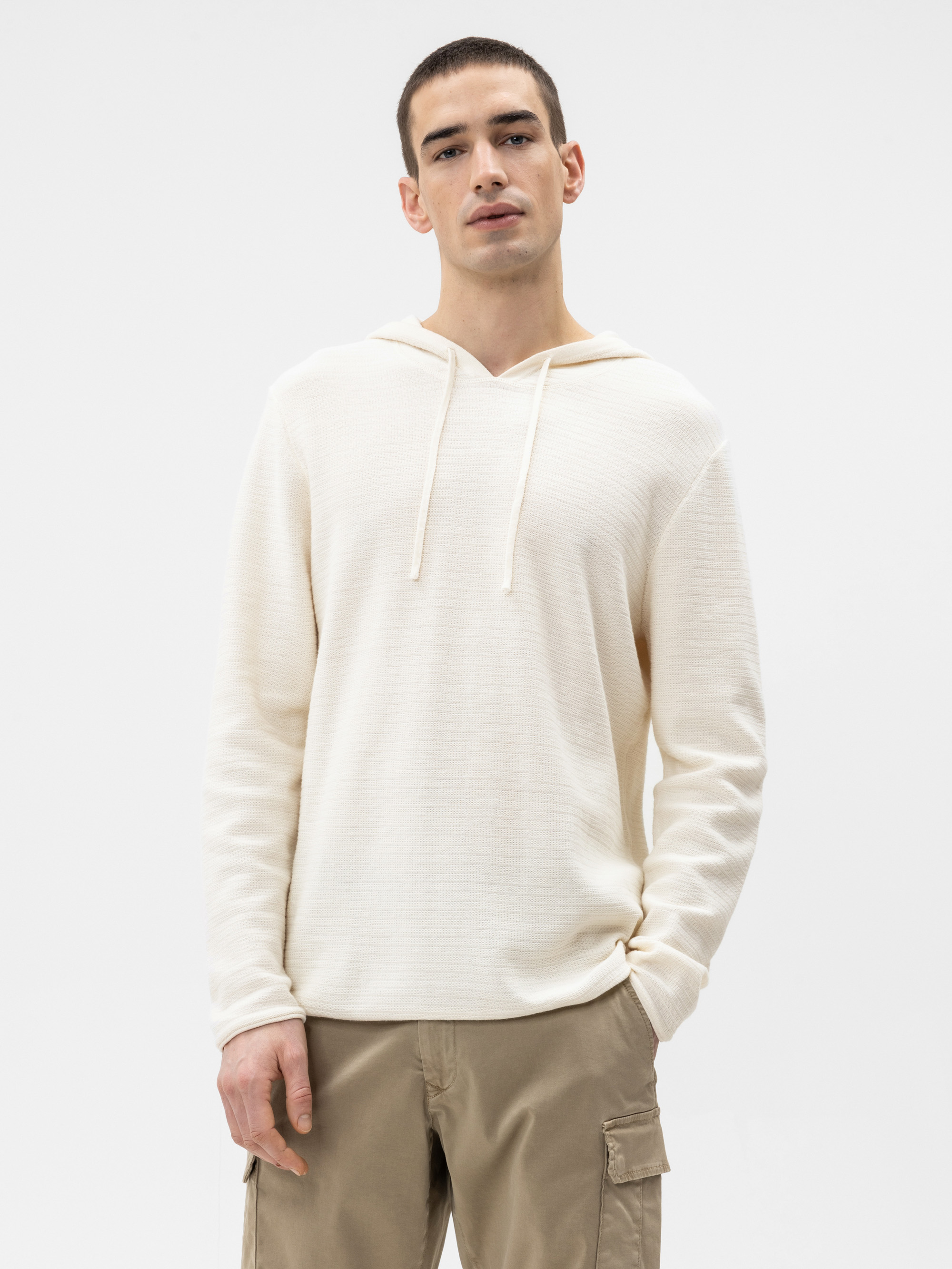 Ein junger Mann, der einen cremefarbenen, langärmeligen Kapuzenpullover mit V-Ausschnitt und Kordelzug sowie eine khakifarbene Cargohose trägt, steht vor einem schlichten weißen Hintergrund.
