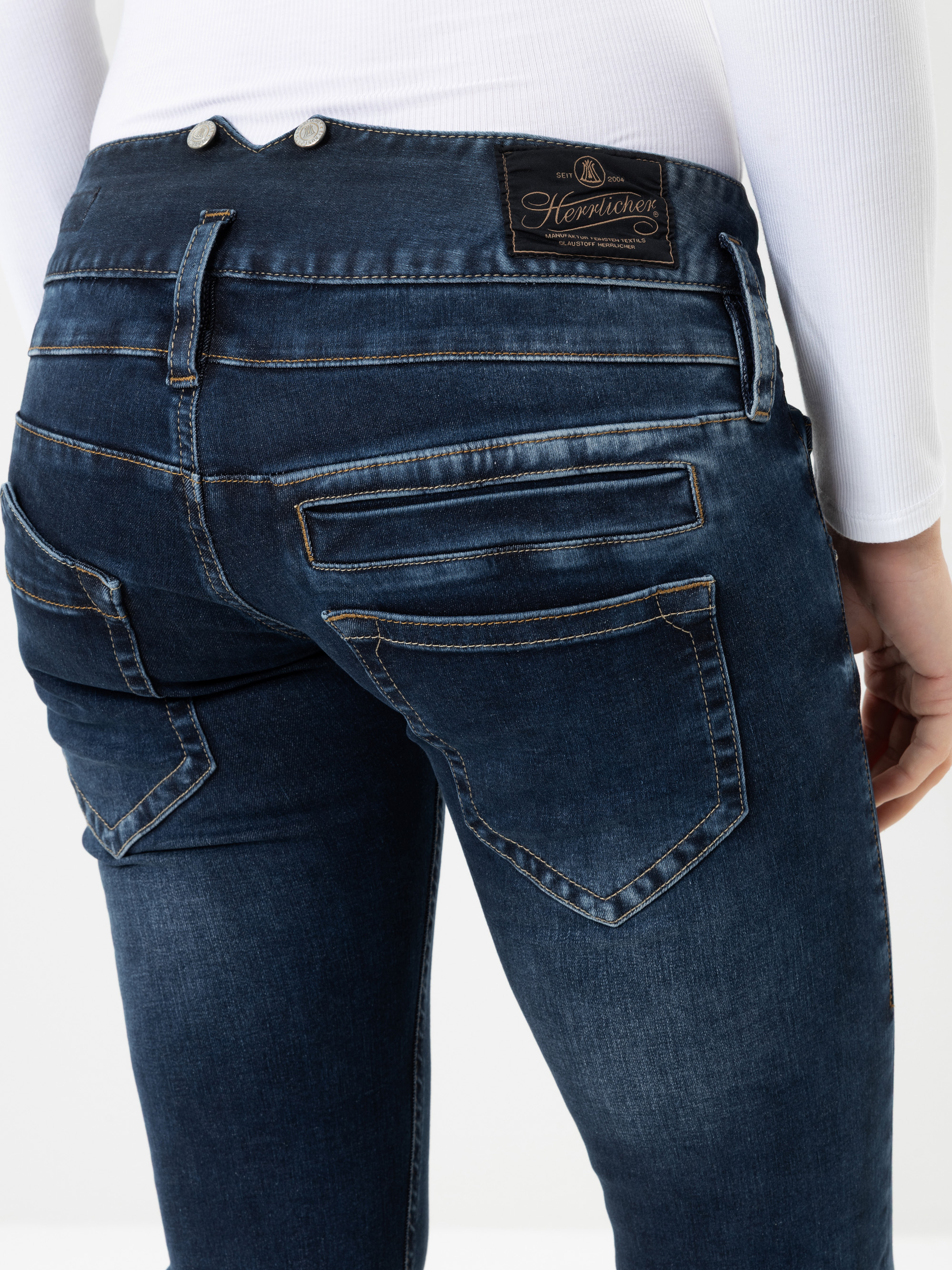 Eine Person, die dunkelblaue Jeans mit mehreren Gesäßtaschen und einem Etikett mit der Aufschrift „Herrlicher“ am Bund trägt, kombiniert mit einem weißen Langarm-Oberteil. Der Fokus des Bildes liegt auf dem unteren Rücken- und dem oberen Obersche