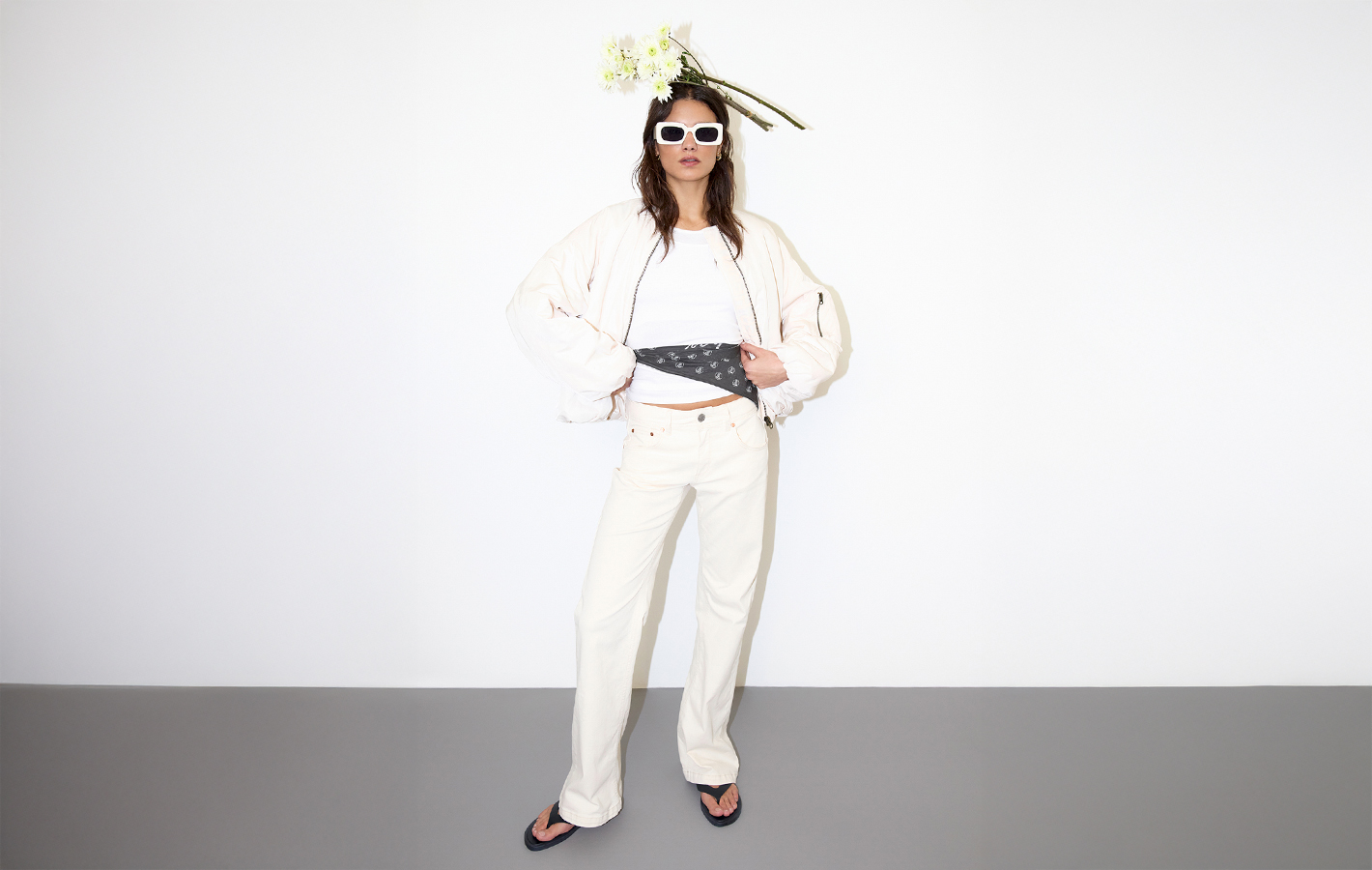 Eine Person mit weißer Sonnenbrille, weißer Jacke, weißer Hose und Flip-Flops steht selbstbewusst da. Sie trägt ein Blumenarrangement auf dem Kopf und hebt ihr Hemd, um ein gemustertes Kopftuch um die Taille zu enthüllen.