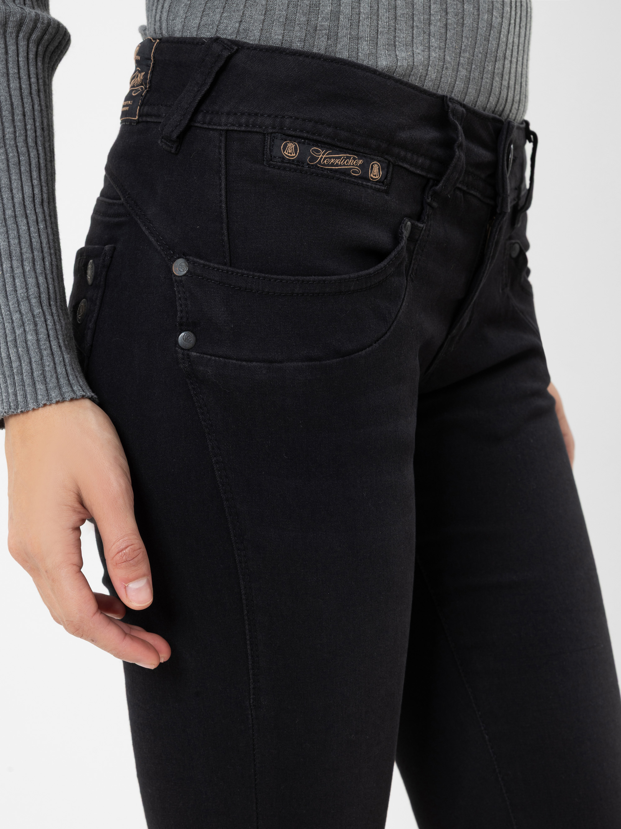 Eine Person, die einen grauen, gerippten Pullover und eine schwarze Jeans mit sichtbaren Knöpfen und Ziernähten trägt; die Abbildung zeigt den Bereich von der Taille bis zur Mitte des Oberschenkels.