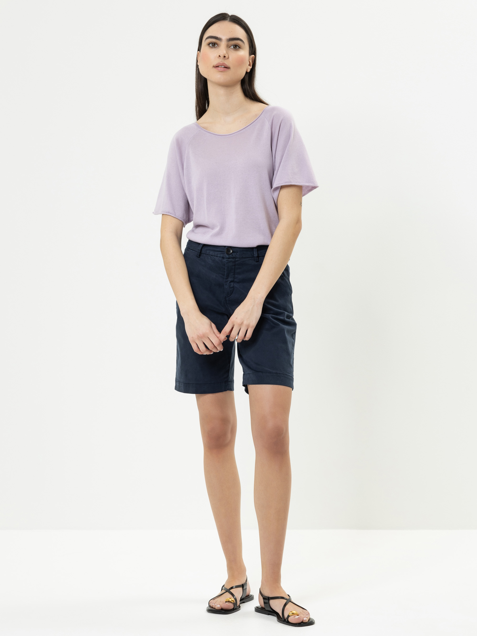 Eine Frau steht vor einem schlichten weißen Hintergrund und trägt ein lockeres, hellviolettes T-Shirt, dunkle marineblaue knielange Shorts und schwarze Sandalen. Sie hat glattes dunkles Haar und einen neutralen Gesichtsausdruck, die Arme liegen entspann