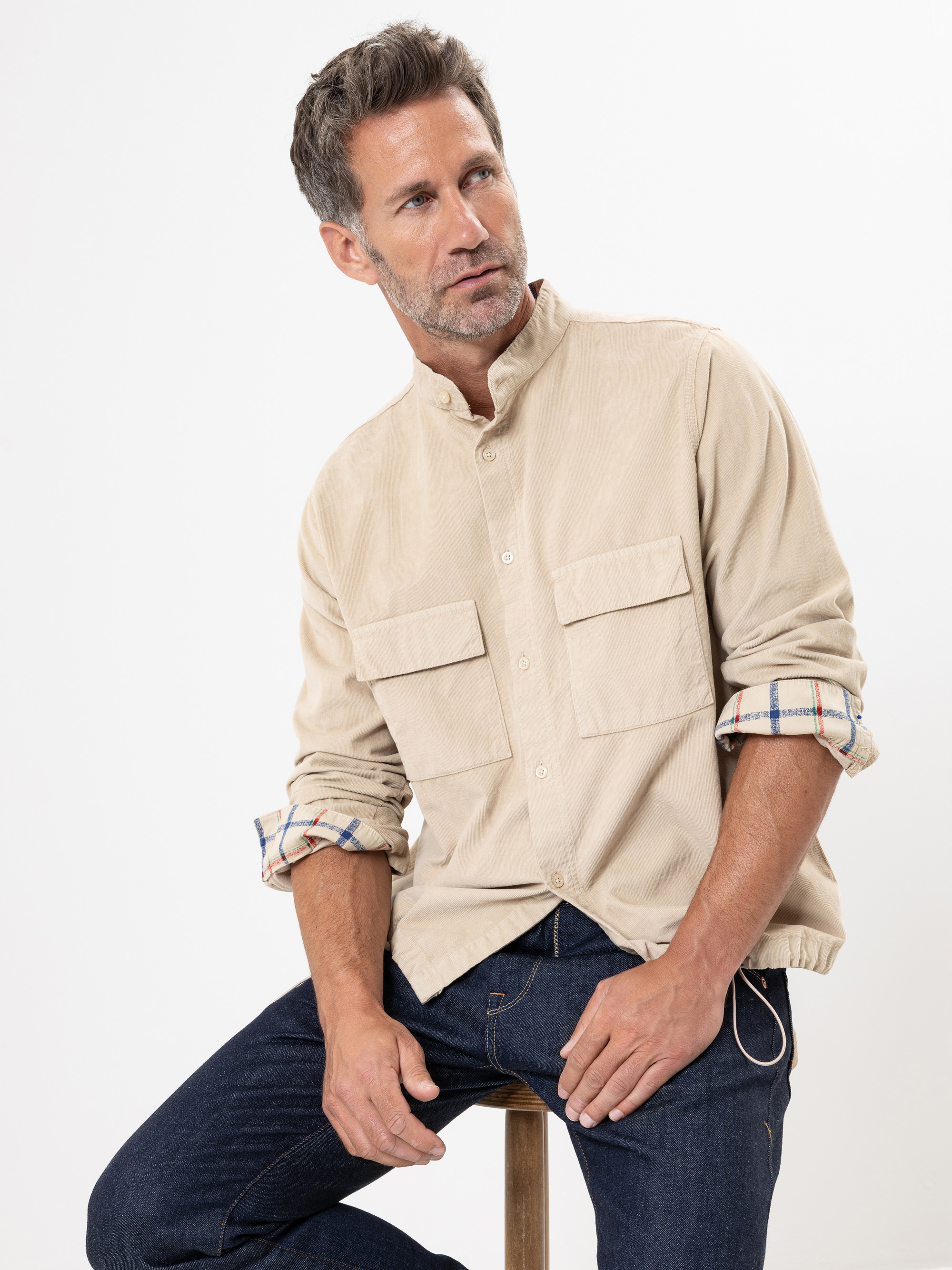 Ein Mann mit kurzen braunen Haaren und gestutztem Bart sitzt auf einem Hocker, trägt ein beiges Button-up-Hemd mit gekrempelten Ärmeln und dunkle Jeans und blickt leicht nach links vor einem schlichten weißen Hintergrund.