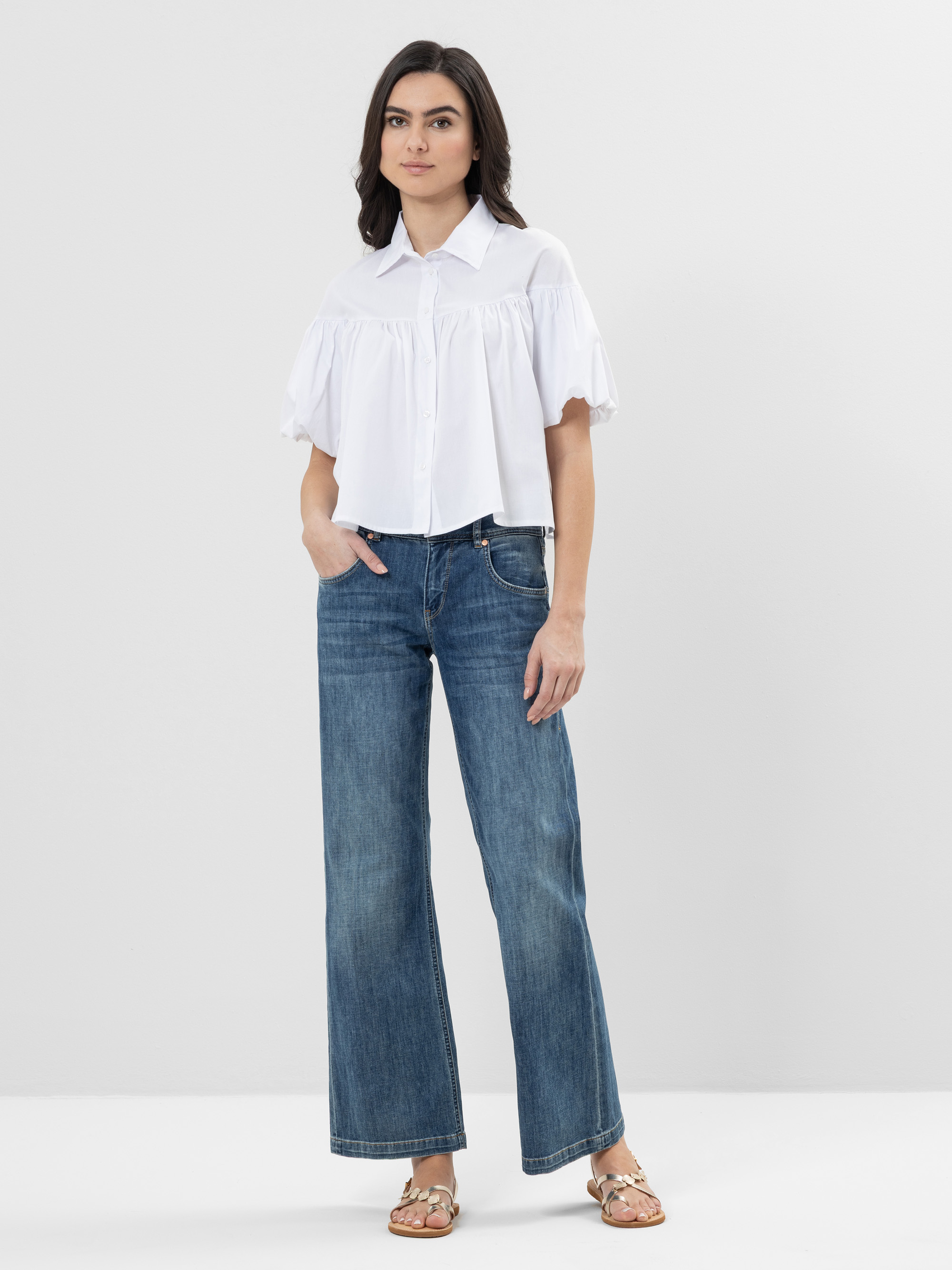 Eine Frau steht vor einem schlichten weißen Hintergrund und trägt eine kurzärmelige weiße Button-up-Bluse, blaue Jeans mit weitem Bein und flache Riemchensandalen. Eine Hand steckt in ihrer Hosentasche und sie blickt mit einem neutralen Blick nach vor