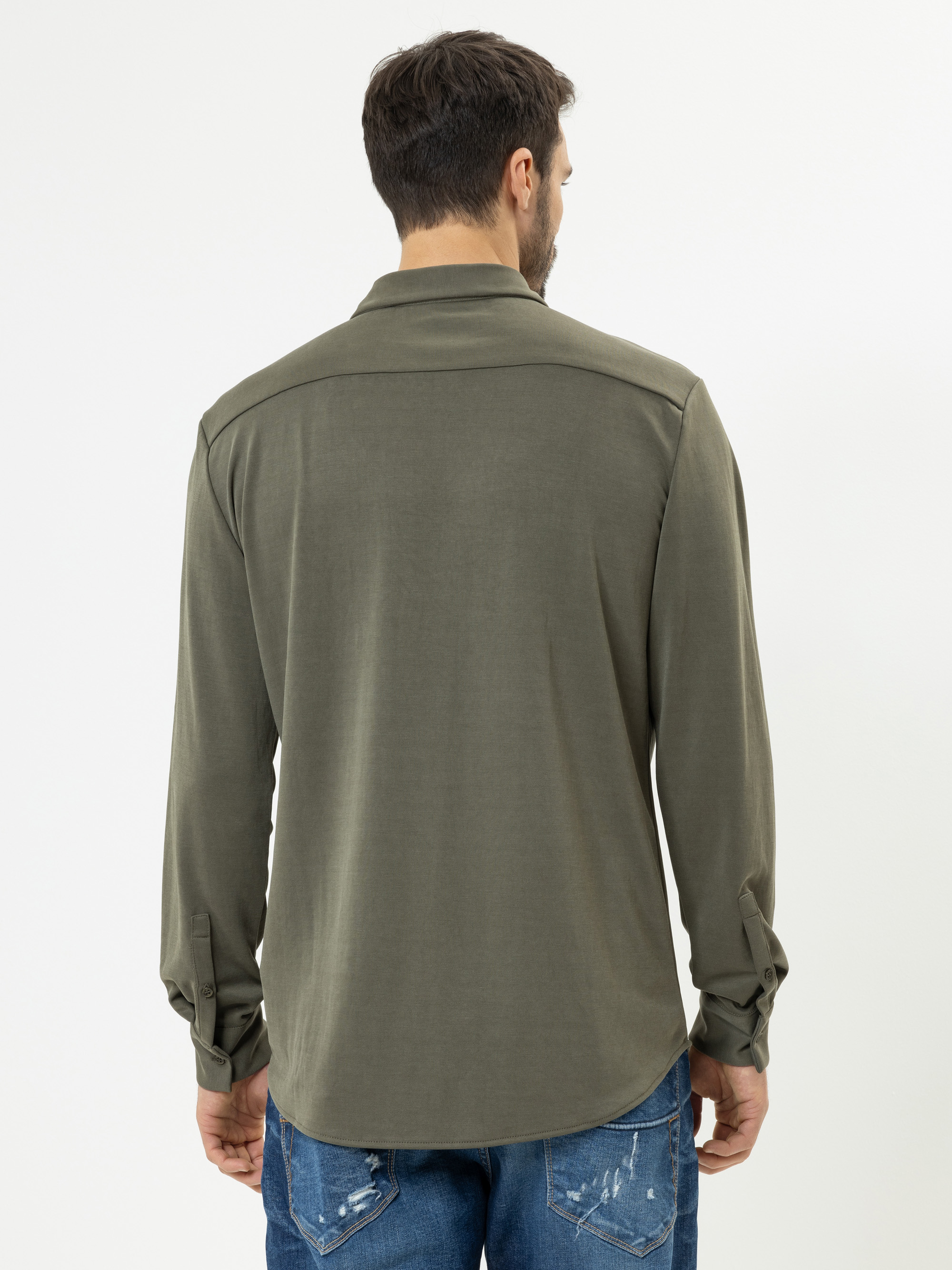 Ein Mann steht mit dem Rücken zur Kamera. Er trägt ein olivgrünes, langärmeliges Hemd und eine blaue, abgewetzte Jeans vor einem schlichten weißen Hintergrund.