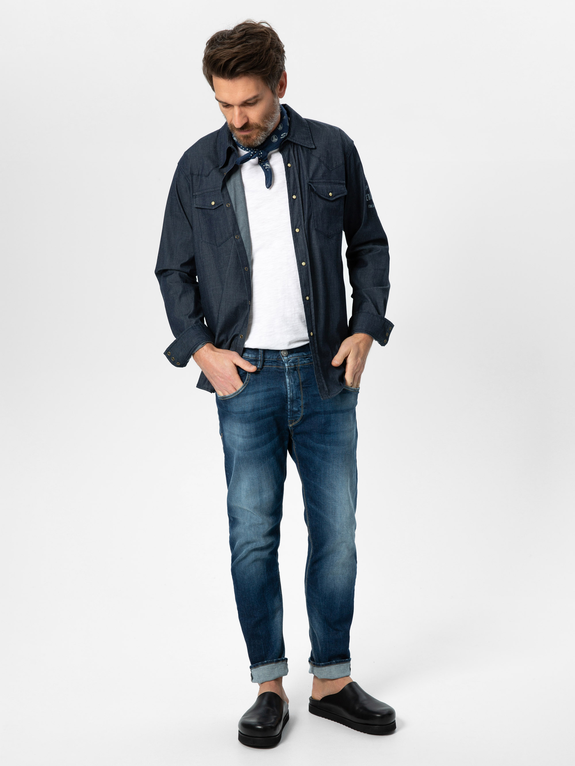 Ein Mann, der ein blaues Button-up-Hemd über einem weißen T-Shirt, blaue Jeans, einen blauen Schal um den Hals und schwarze Slipper trägt, steht mit den Händen in den Taschen und blickt auf einen schlichten weißen Hintergrund.