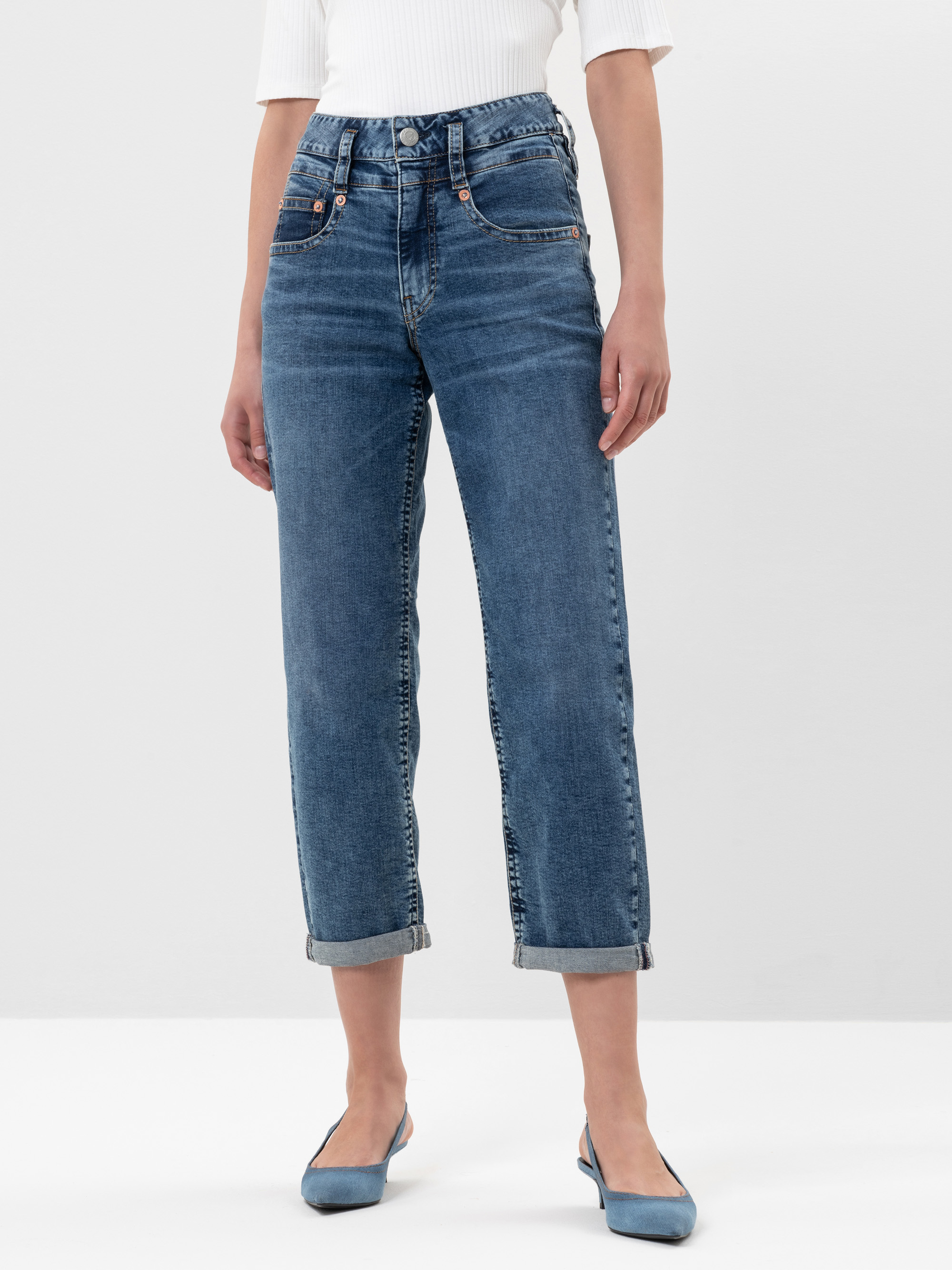 Eine Person, die hochgekrempelte blaue Jeans, ein weißes geripptes Oberteil mit ellbogenlangen Ärmeln und blaue, spitz zulaufende Wohnungen trägt, steht vor einem schlichten weißen Hintergrund.