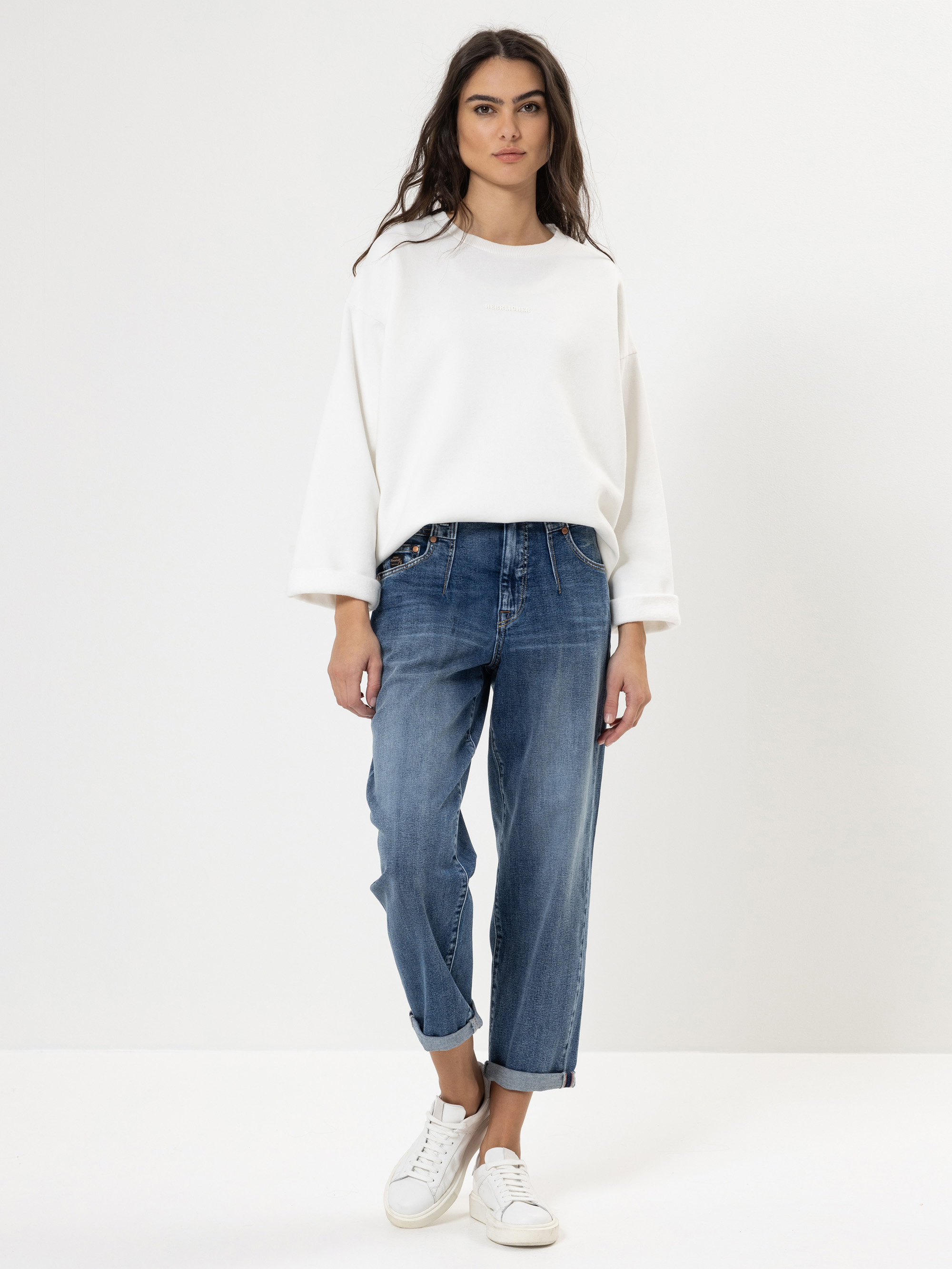 Eine Frau steht vor einem schlichten weißen Hintergrund und trägt ein weißes übergroßes Sweatshirt, locker sitzende Bluejeans mit aufgerollten Bündchen und weiße Turnschuhe. Sie hat langes dunkles Haar und einen neutralen Gesichtsausdruck.