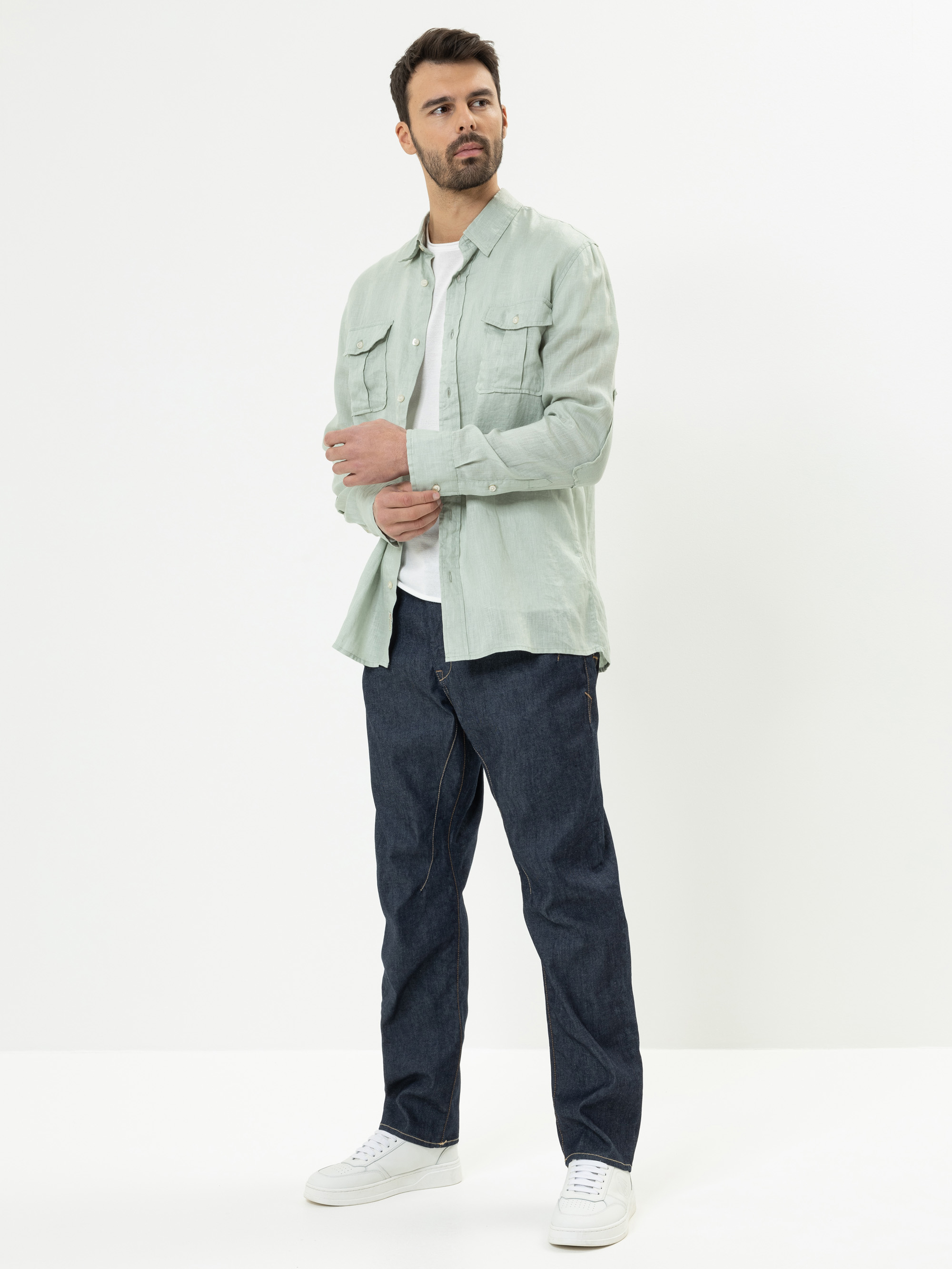 Ein Mann steht vor einem schlichten weißen Hintergrund und trägt ein hellgrünes Button-up-Hemd über einem weißen T-Shirt, dunkelblaue Jeans und weiße Turnschuhe. Er blickt zur Seite, während er mit einer Hand sein Hemd zurechtrückt.