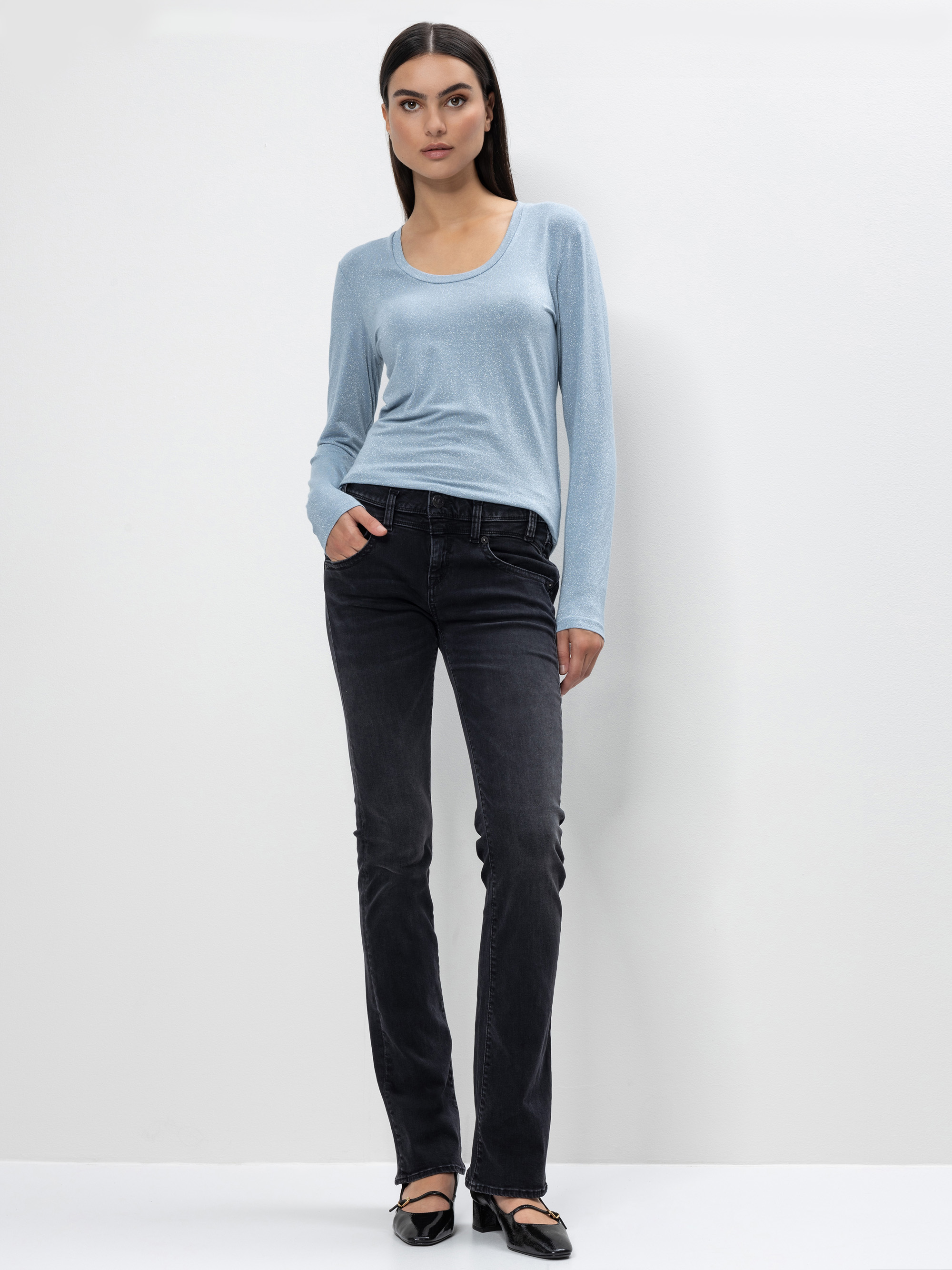 Eine Frau mit langen dunklen Haaren steht vor einer schlichten weißen Wand. Sie trägt ein hellblaues langärmeliges Oberteil, schwarze Jeans und schwarze Flats, eine Hand in der Tasche und einen neutralen Gesichtsausdruck.