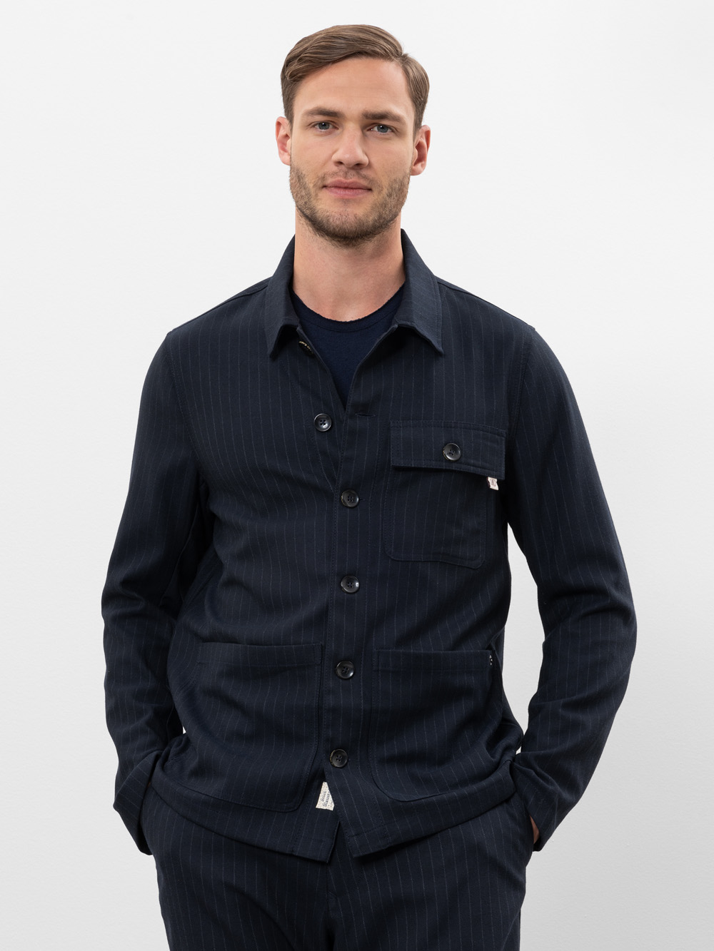 Ein Mann mit kurzen hellbraunen Haaren und gestutztem Bart trägt eine dunkle, marineblaue Jacke mit Nadelstreifen und eine dazu passende Hose und steht mit den Händen in den Taschen vor einem schlichten weißen Hintergrund.
