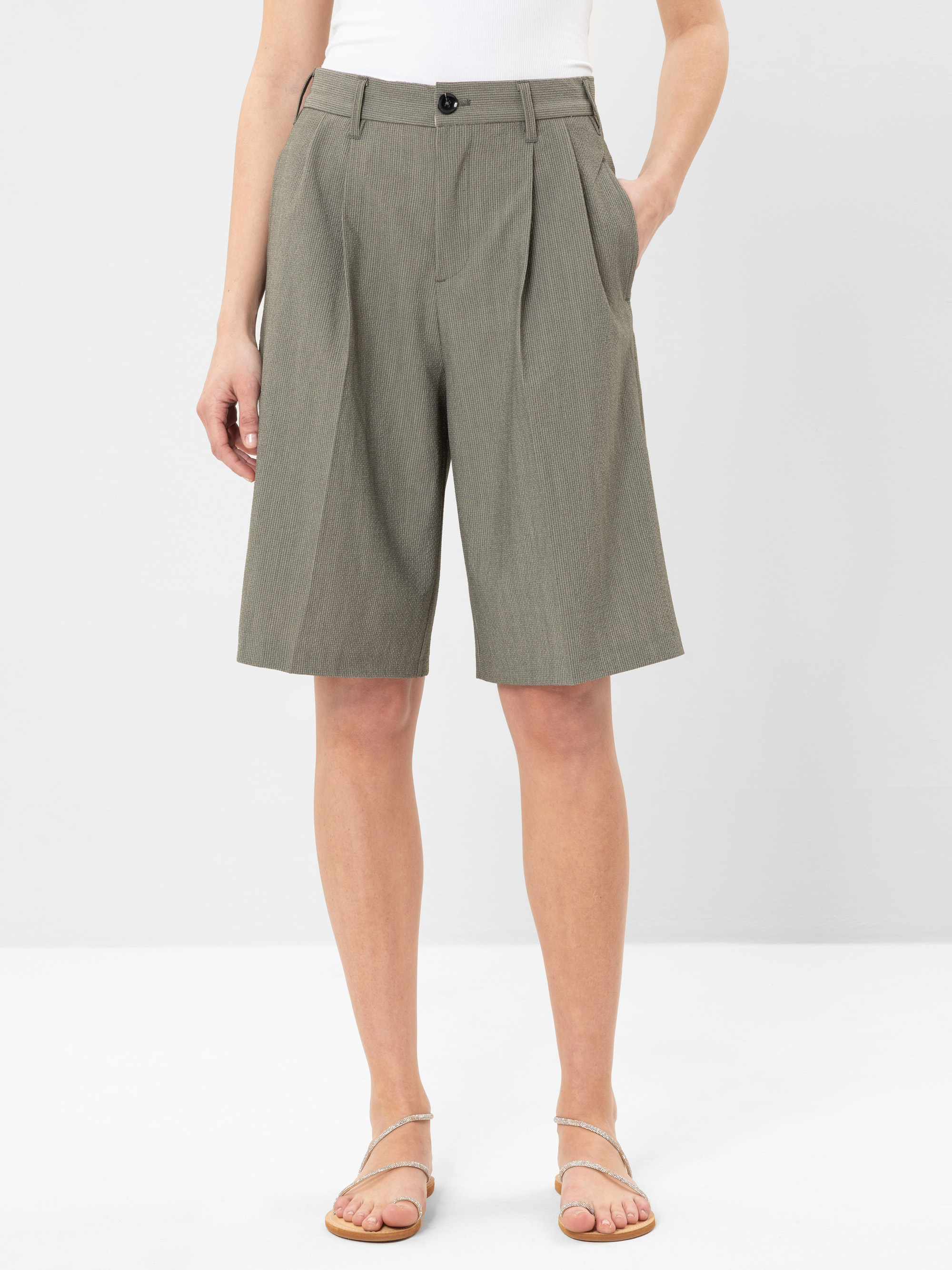 Eine Person, die ein weißes Oberteil und eine knielange, hochtaillierte, in Falten gelegte graue Hose mit Taschen und einem schwarzen Knopf trägt, dazu flache Riemchensandalen, steht vor einem einfarbig weißen Hintergrund.