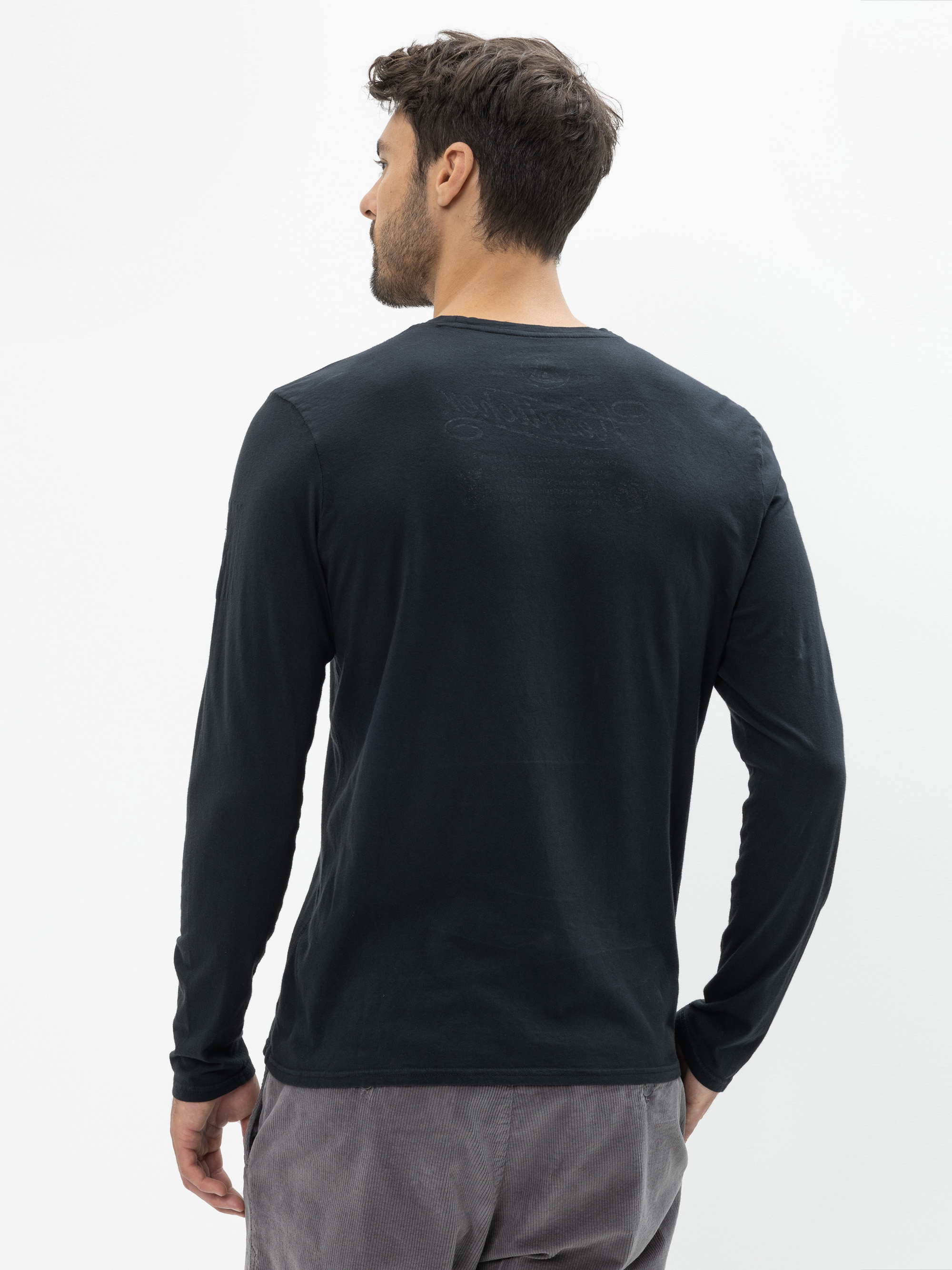 Ein Mann mit kurzen braunen Haaren steht mit dem Rücken zur Kamera, trägt ein langärmeliges schwarzes Hemd und eine graue Hose vor einem schlichten weißen Hintergrund.