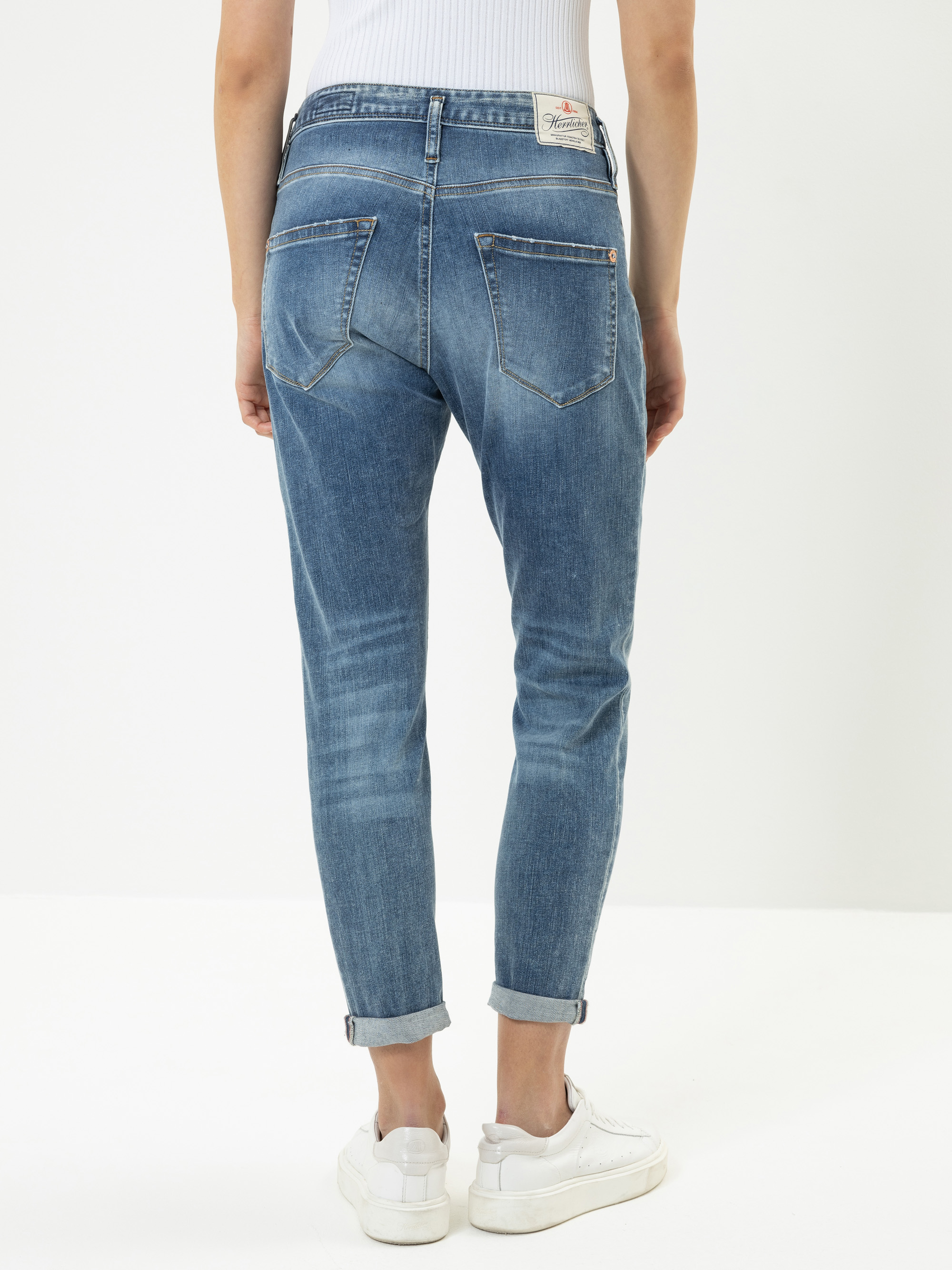 Eine Person in hochgekrempelten Bluejeans und weißen Turnschuhen steht mit dem Gesicht zur Kamera vor einem schlichten weißen Hintergrund. Die Jeans hat sichtbare Nähte, Gesäßtaschen und ein Markenetikett am Bund.