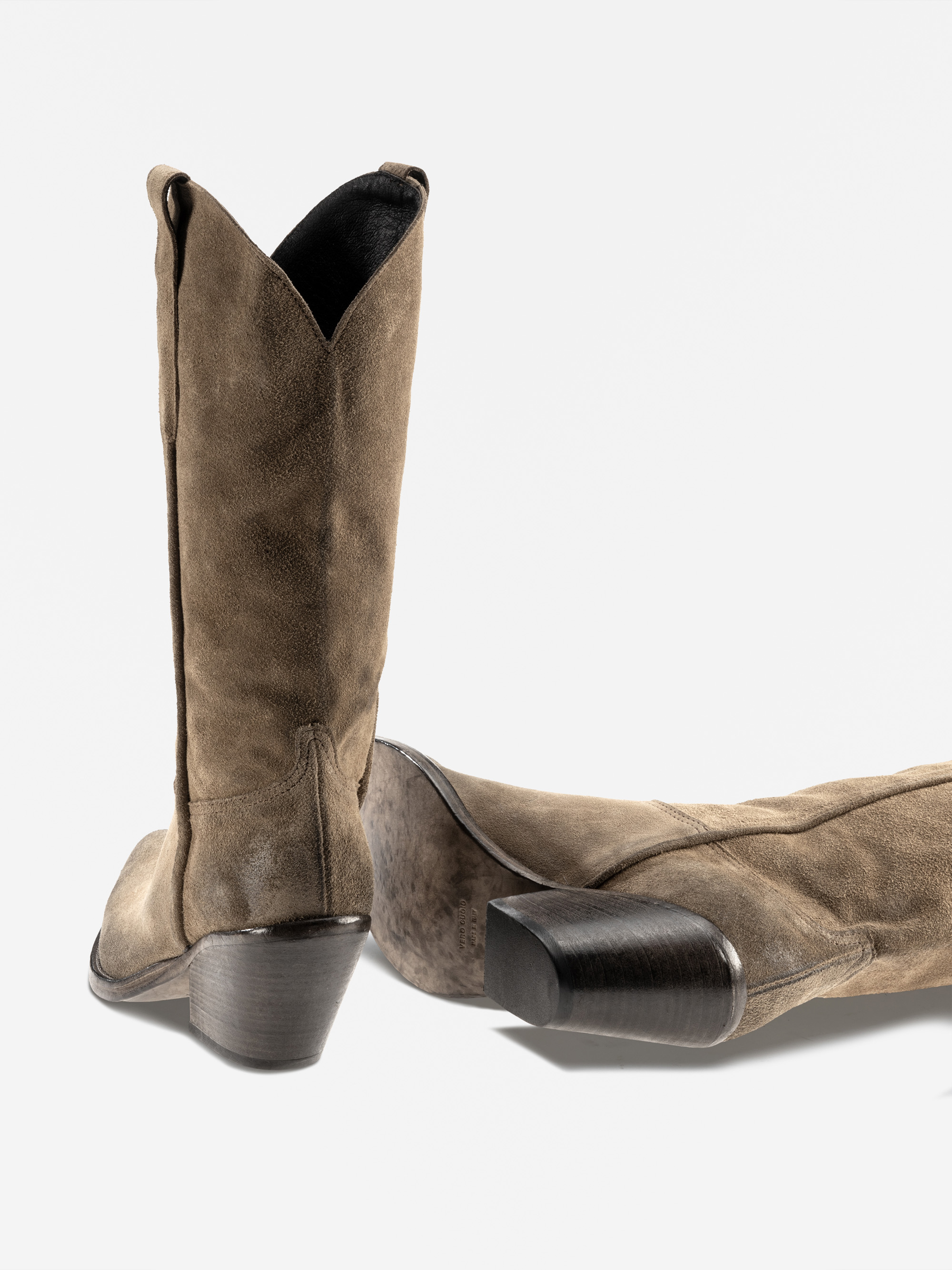 Ein Paar braune Cowboystiefel aus Wildleder mit spitzen Zehen und gestapelten Holzabsätzen, wobei ein Stiefel aufrecht steht und der andere auf der Seite liegt, auf weißem Hintergrund.