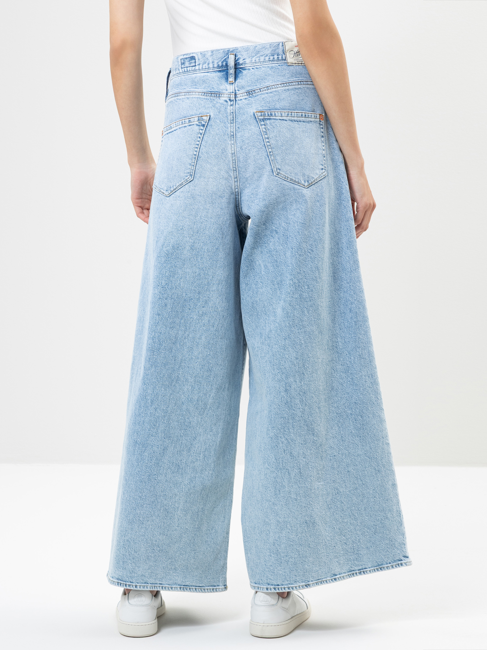 Eine Person, die hellblaue Jeans mit weitem Bein und weiße Turnschuhe trägt, steht mit dem Rücken zur Kamera vor einem schlichten weißen Hintergrund.