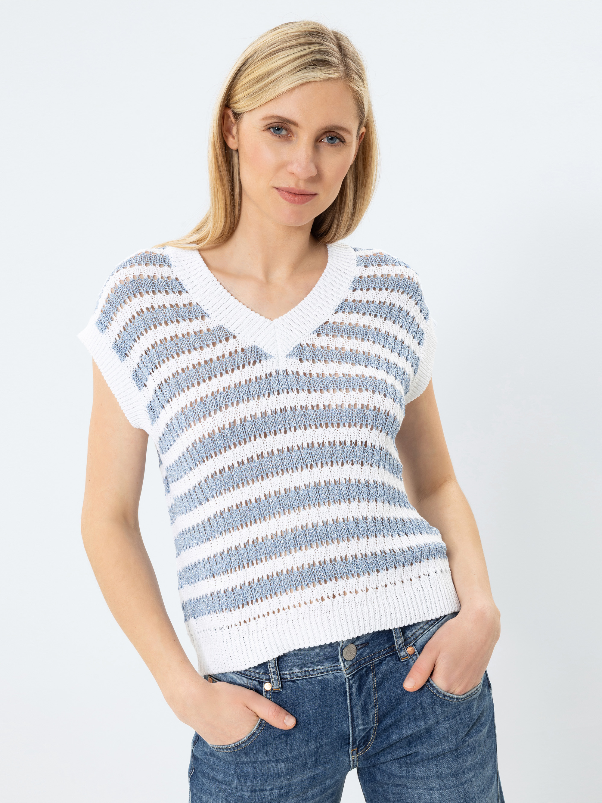 Eine Frau mit blondem Haar trägt einen weiß-blau gestreiften, kurzärmeligen Strickpullover und blaue Jeans und steht mit einer Hand in der Tasche vor einem schlichten, hellen Hintergrund.