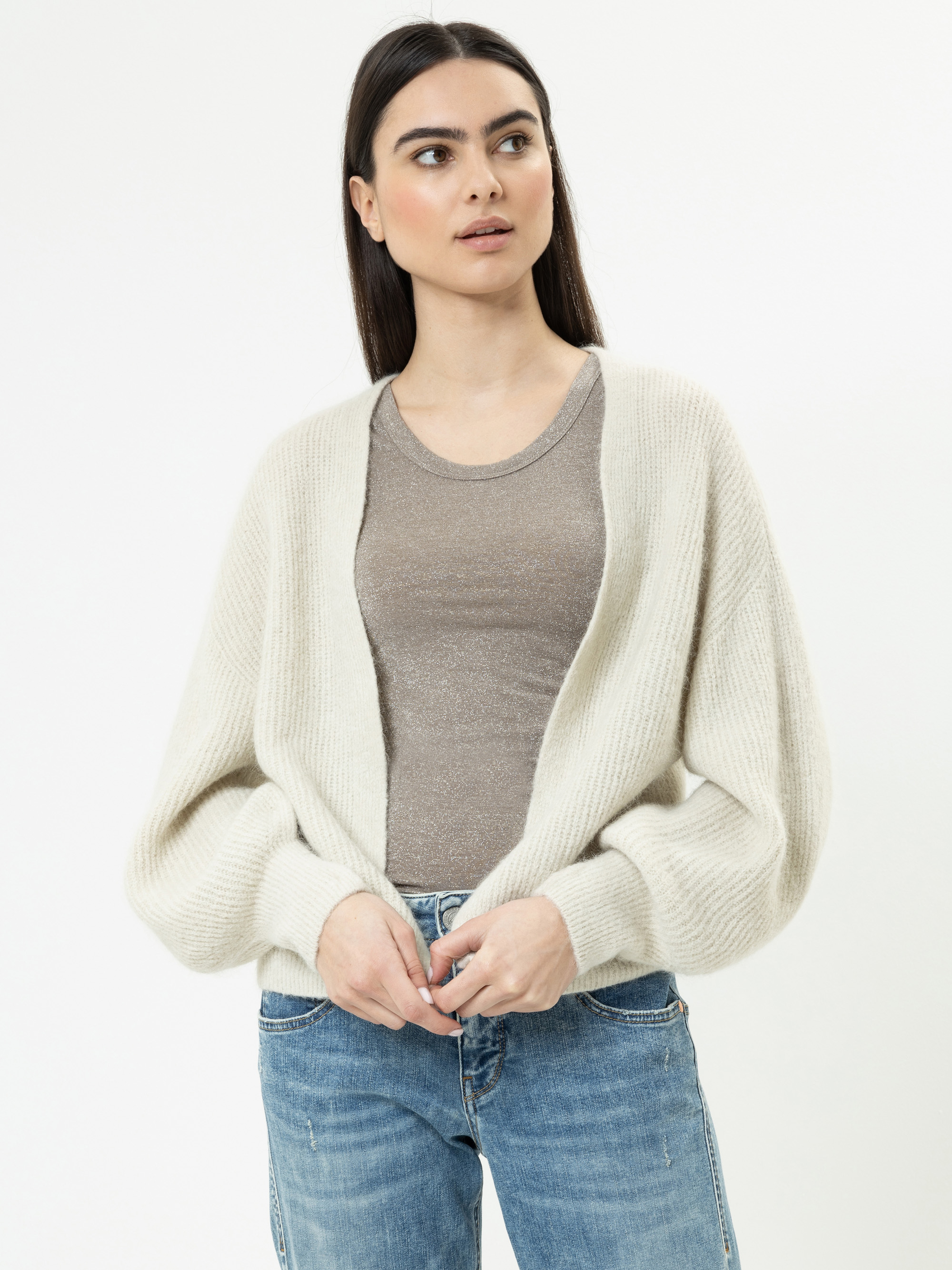 Eine Frau mit langen dunklen Haaren trägt eine cremefarbene Strickjacke über einem beigen Oberteil und blaue Jeans. Sie steht vor einem schlichten weißen Hintergrund und schaut leicht zur Seite.