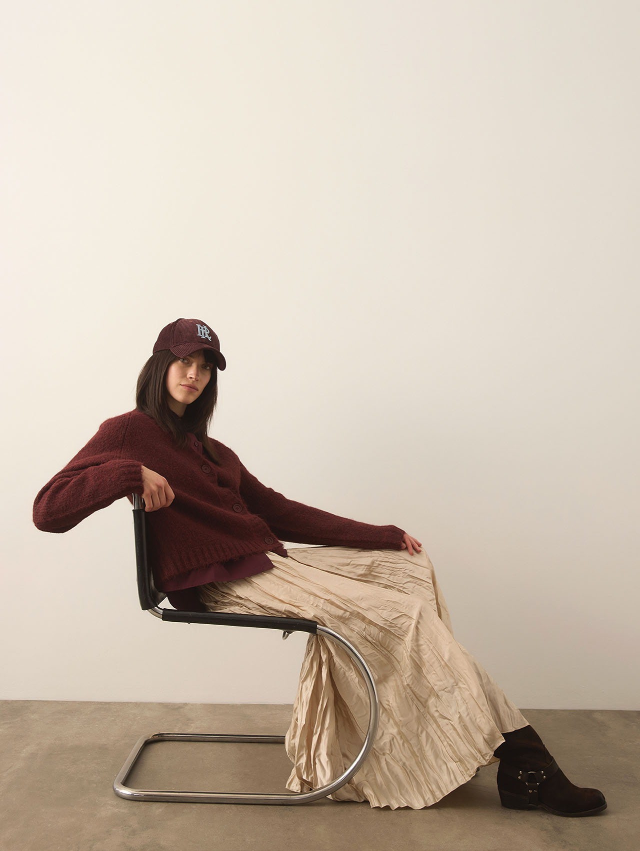 Eine Frau sitzt lässig auf einem modernen Metallstuhl, trägt einen kastanienbraunen Pullover, eine passende Mütze, einen langen beigen Faltenrock und dunkle Stiefel vor einer schlichten hellen Wand und einem Betonboden.