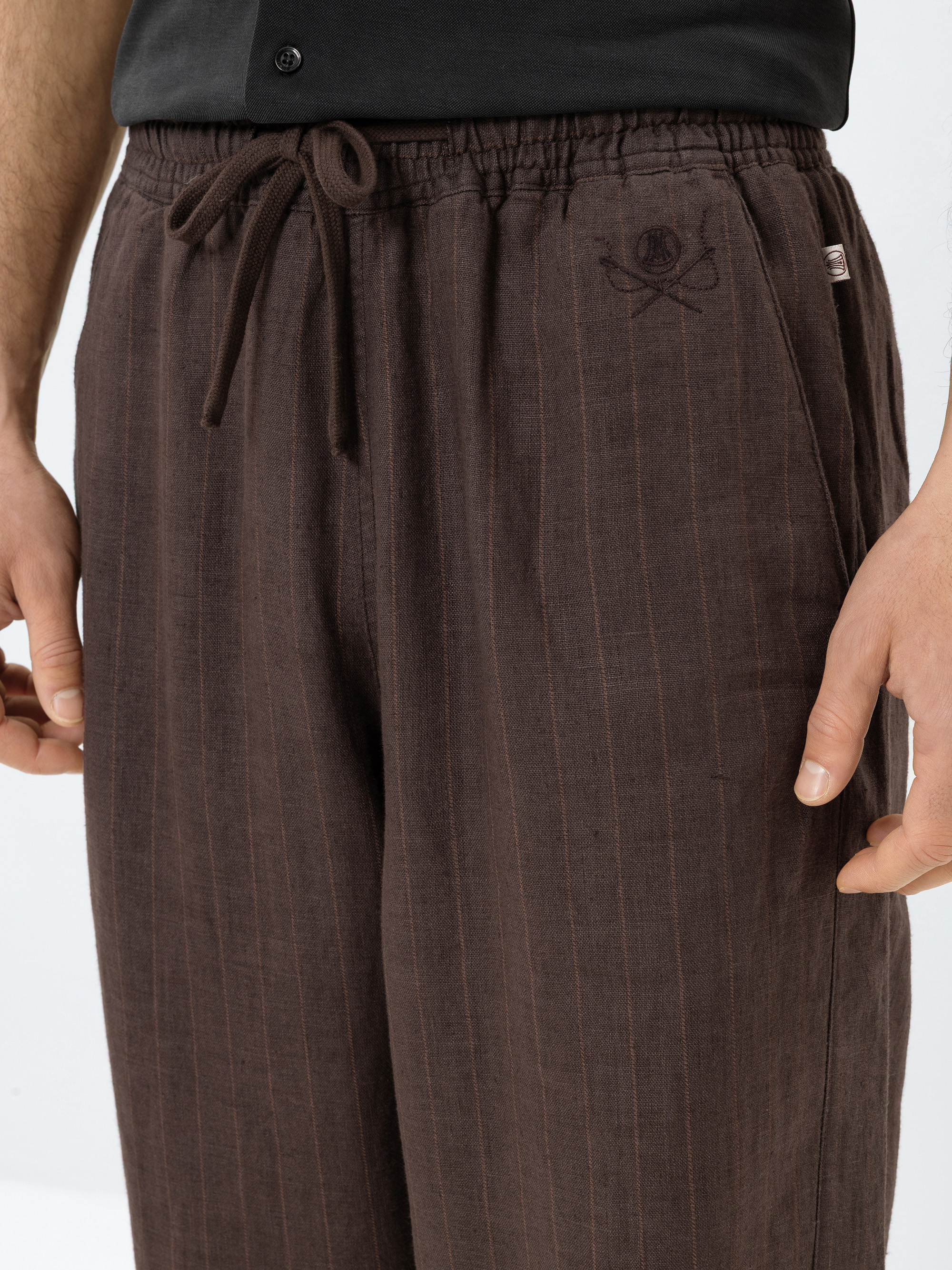 Eine Person, die eine locker sitzende braune Nadelstreifenhose mit Kordelzug in der Taille und einem kleinen gestickten Emblem in der Nähe der linken Tasche trägt, steht mit entspannt an der Seite gehaltenen Händen.