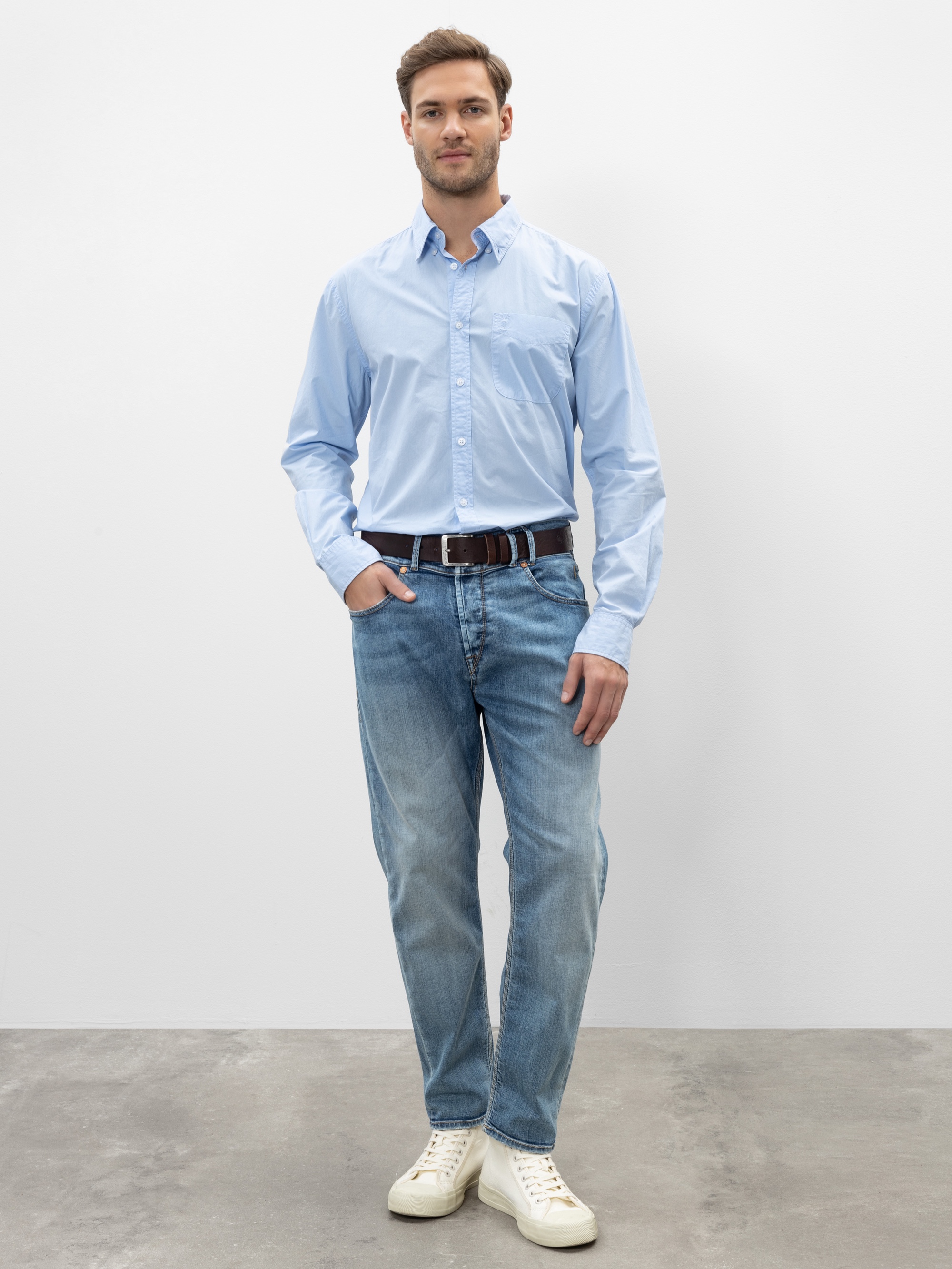 Ein Mann steht vor einem schlichten weißen Hintergrund, trägt ein hellblaues Button-up-Hemd, verblichene Bluejeans, einen braunen Gürtel und weiße Turnschuhe und hat eine Hand in der Tasche.
