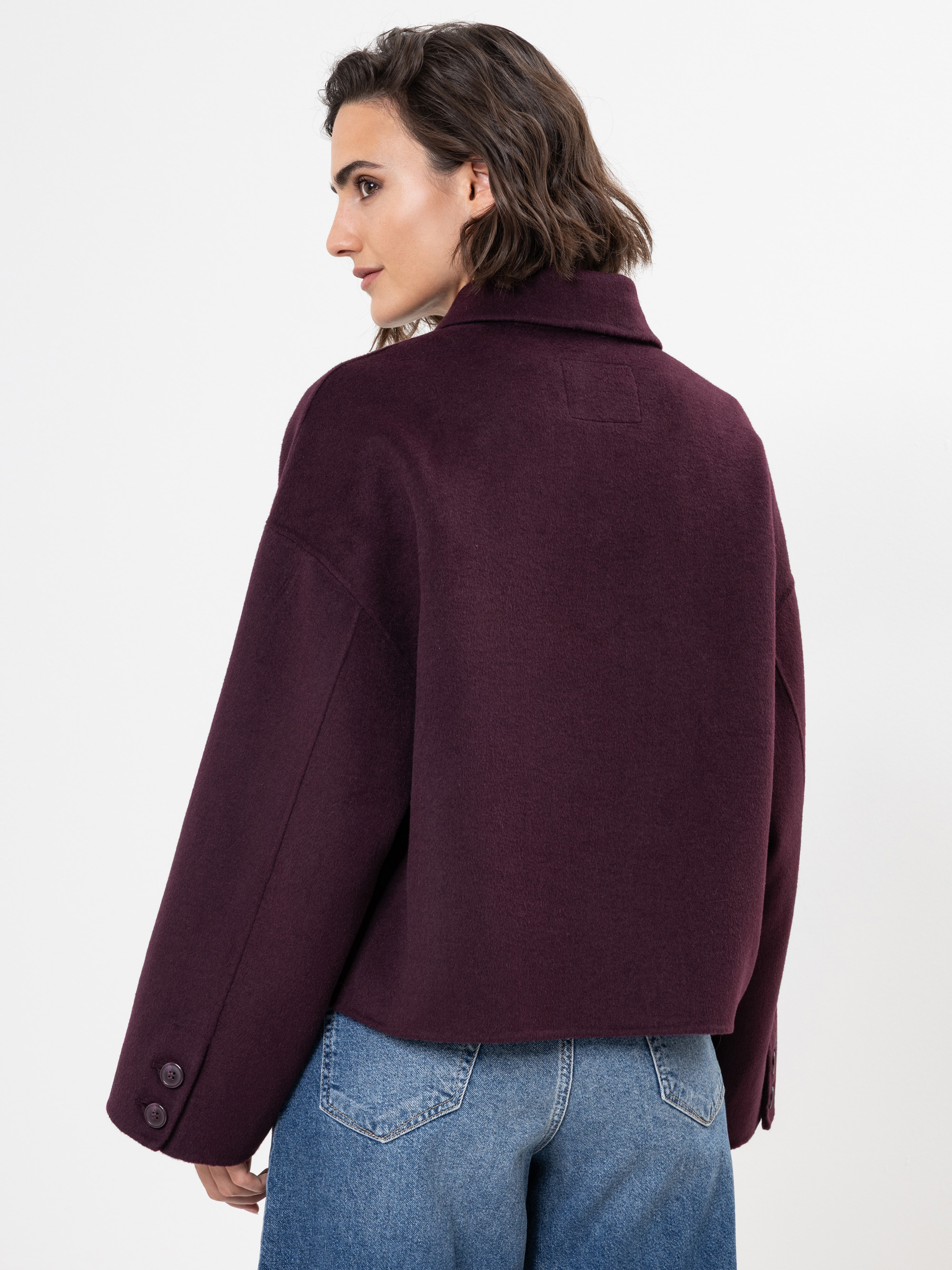 Eine Frau mit kurzen braunen Haaren, die einen locker sitzenden, dunkelvioletten Mantel und blaue Jeans trägt, steht mit leicht zur Seite gedrehtem Kopf vor einem schlichten weißen Hintergrund.