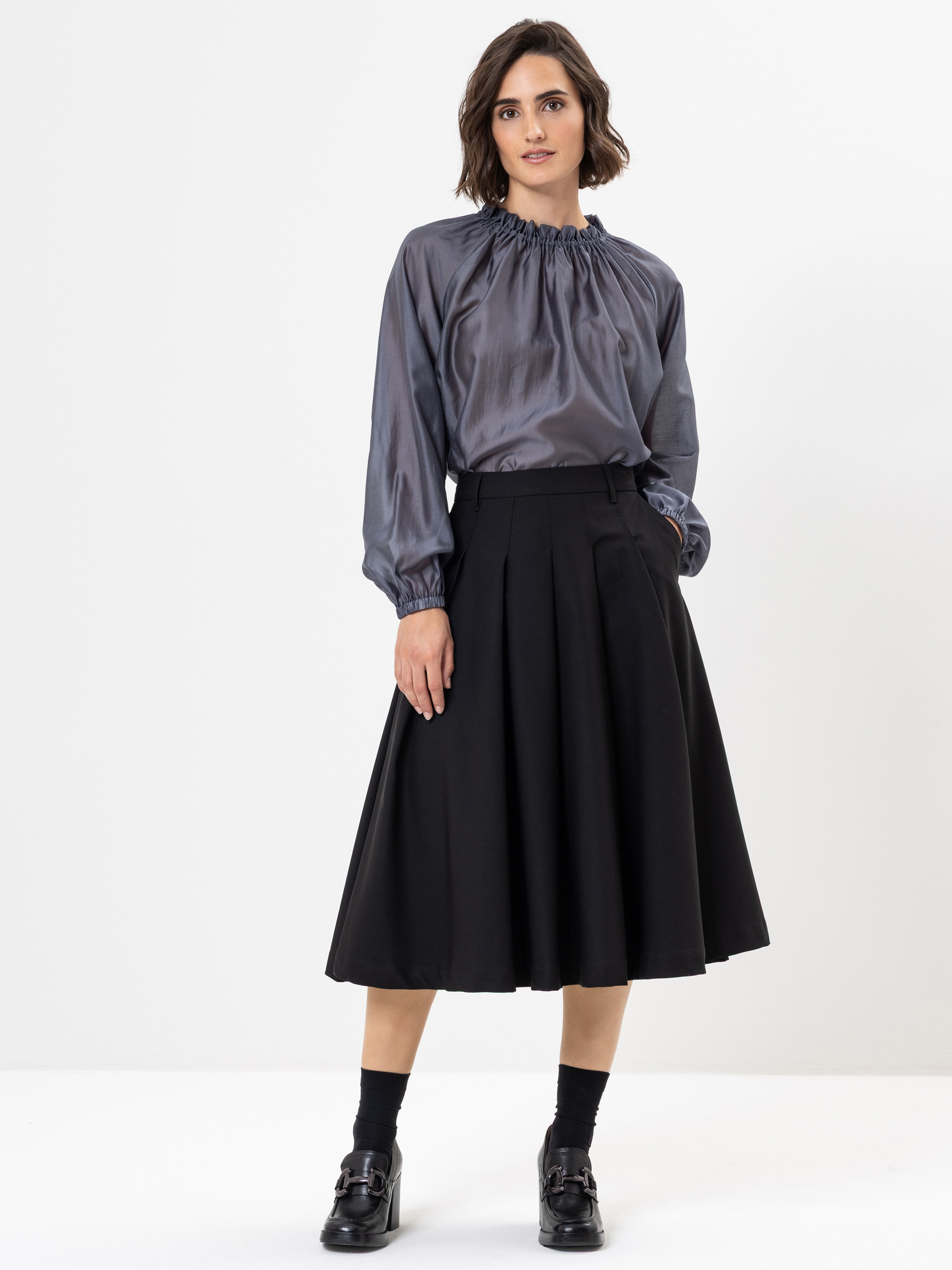 Eine Frau steht vor einem schlichten weißen Hintergrund und trägt eine glänzende graue, langärmelige Bluse mit gerafftem Ausschnitt, einen schwarzen Midirock mit hoher Taille, schwarze Socken und schwarze Schuhe.