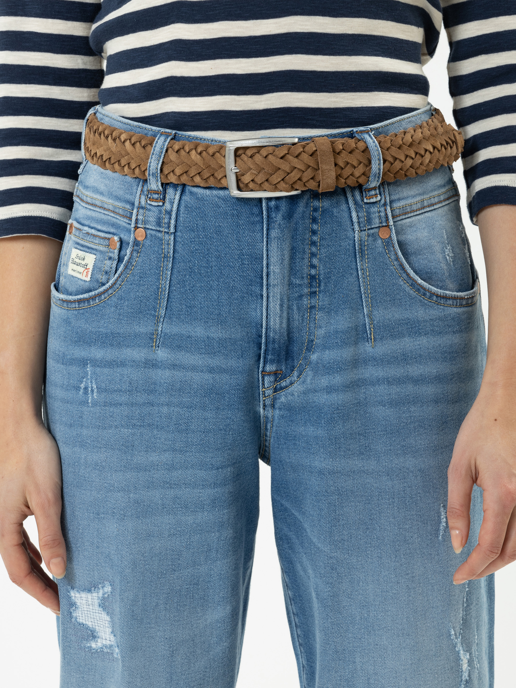 Eine Person, die ein marineblau-weiß gestreiftes Hemd, blaue Jeans mit leichten Gebrauchsspuren und einen braunen Flechtgürtel trägt. Das Foto ist so beschnitten, dass nur der Torso und die Oberschenkel zu sehen sind.