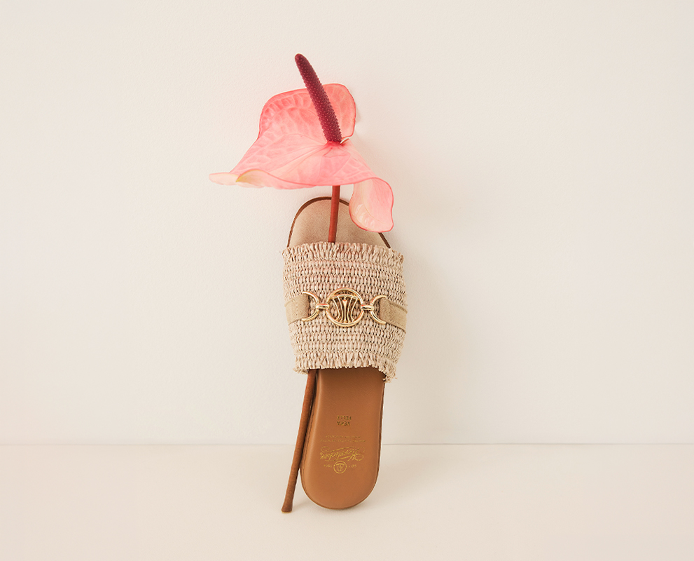 Eine beigefarbene, geflochtene Pantolette mit einem goldenen Schmuckemblem ist aufrecht an eine Wand gelehnt. Im Inneren der Sandale befindet sich eine rosafarbene Anthurienblüte, deren Stiel aus der Sandale herausragt.