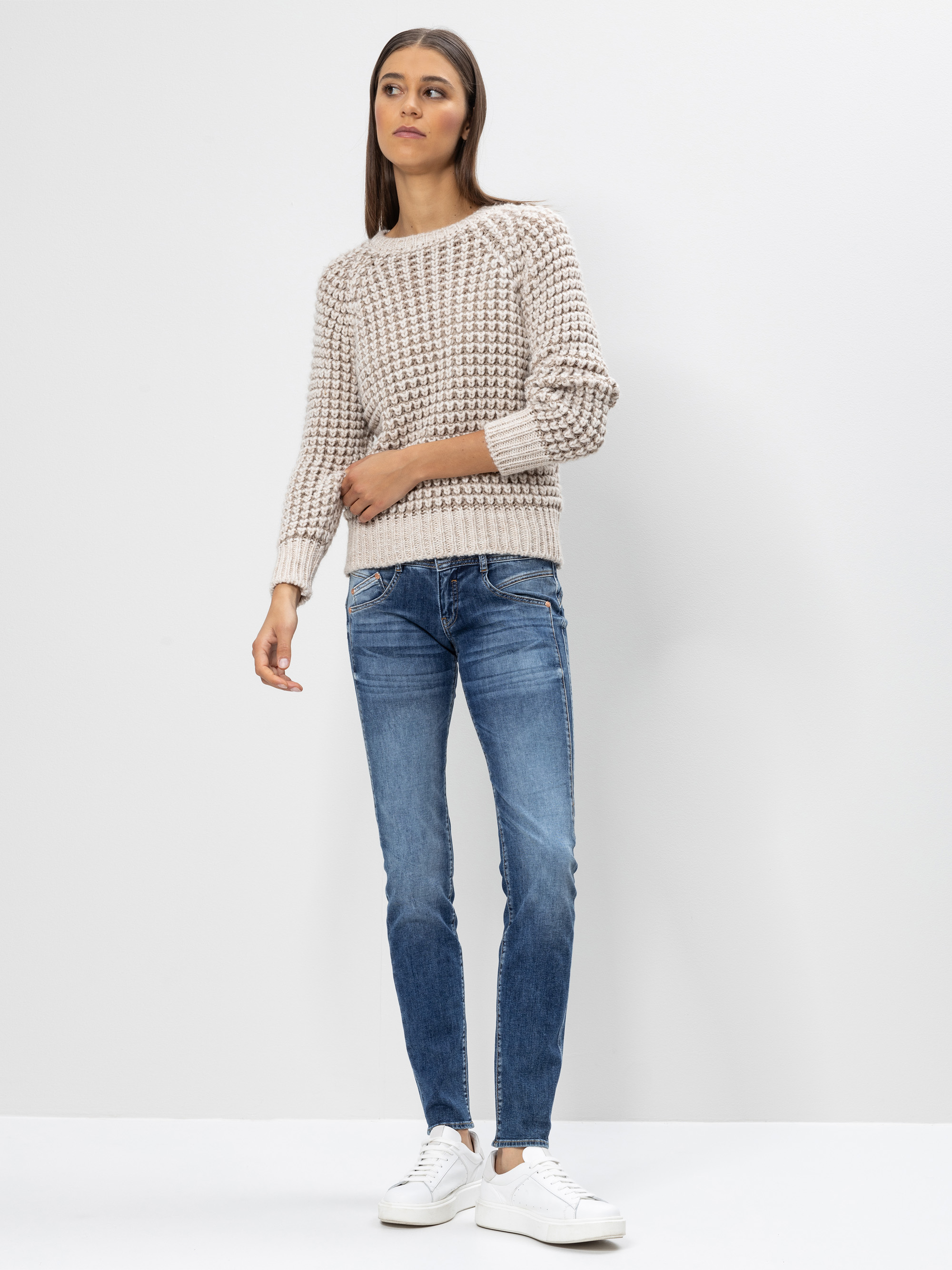 Eine Frau steht vor einem schlichten weißen Hintergrund und trägt einen beigen Strickpullover, blaue Skinny Jeans und weiße Turnschuhe. Sie hat langes, glattes braunes Haar und einen neutralen Gesichtsausdruck.