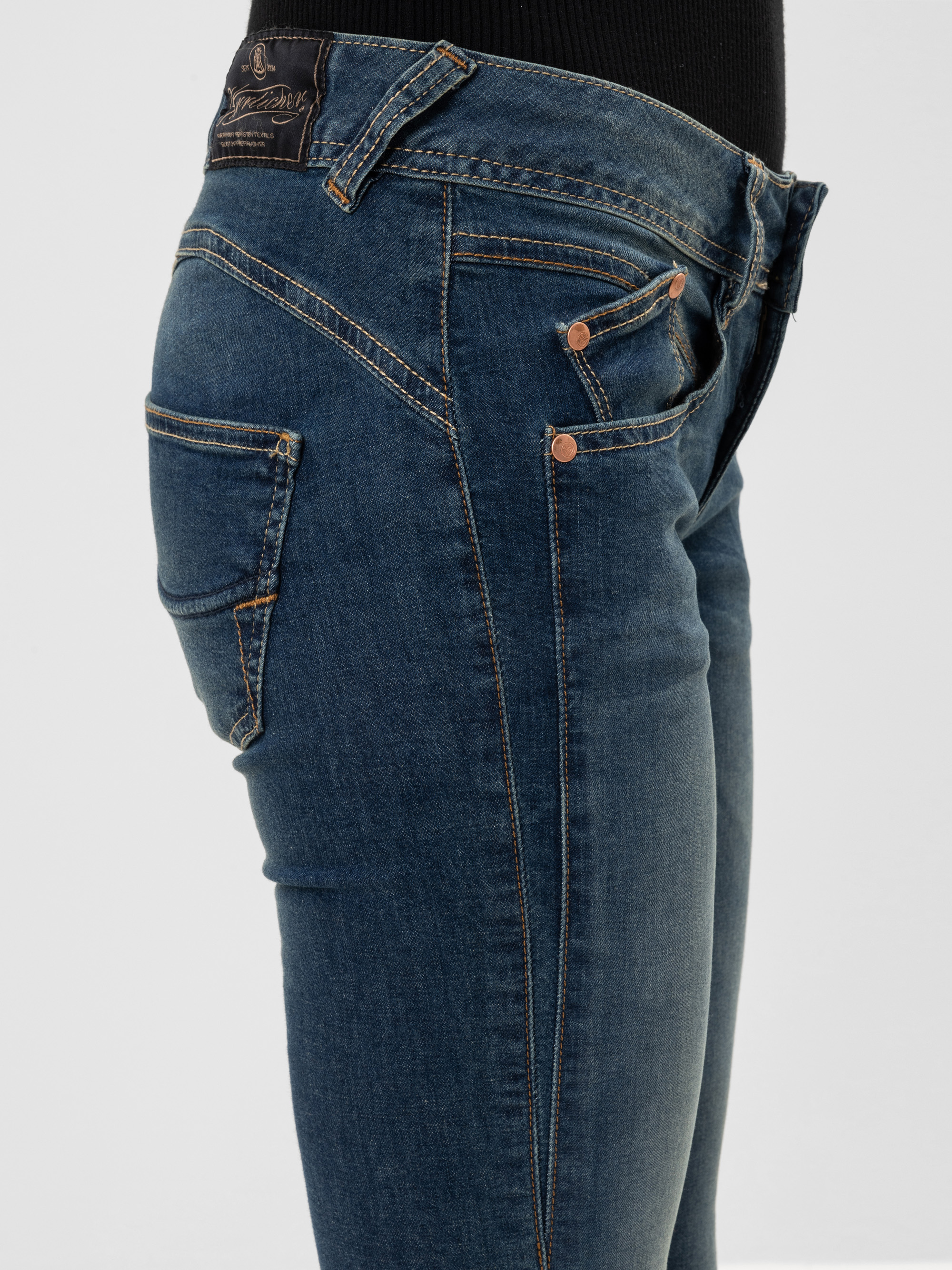 Eine Person, die eine blaue Jeans und ein schwarzes Oberteil trägt, von der Taille abwärts und von der Seite gesehen vor einem schlichten hellen Hintergrund.