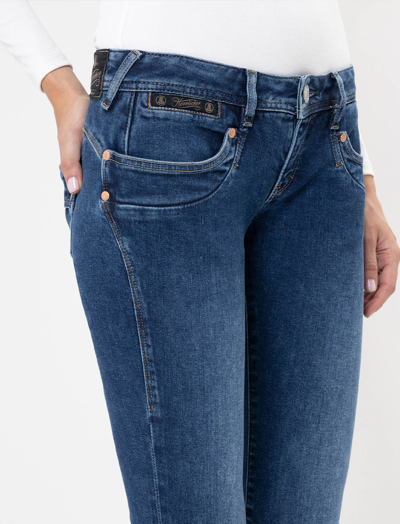 Eine Person, die eine blaue Jeans und ein weißes langärmeliges Oberteil trägt, mit einer Hand in der Tasche, wird von der Mitte des Oberkörpers bis zu den Oberschenkeln vor einem unifarbenen Hintergrund gezeigt.